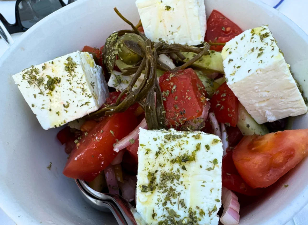 Bowl of Greek salad with feta, tomatoes, cucumber, onions, and herbs on an outdoor table.