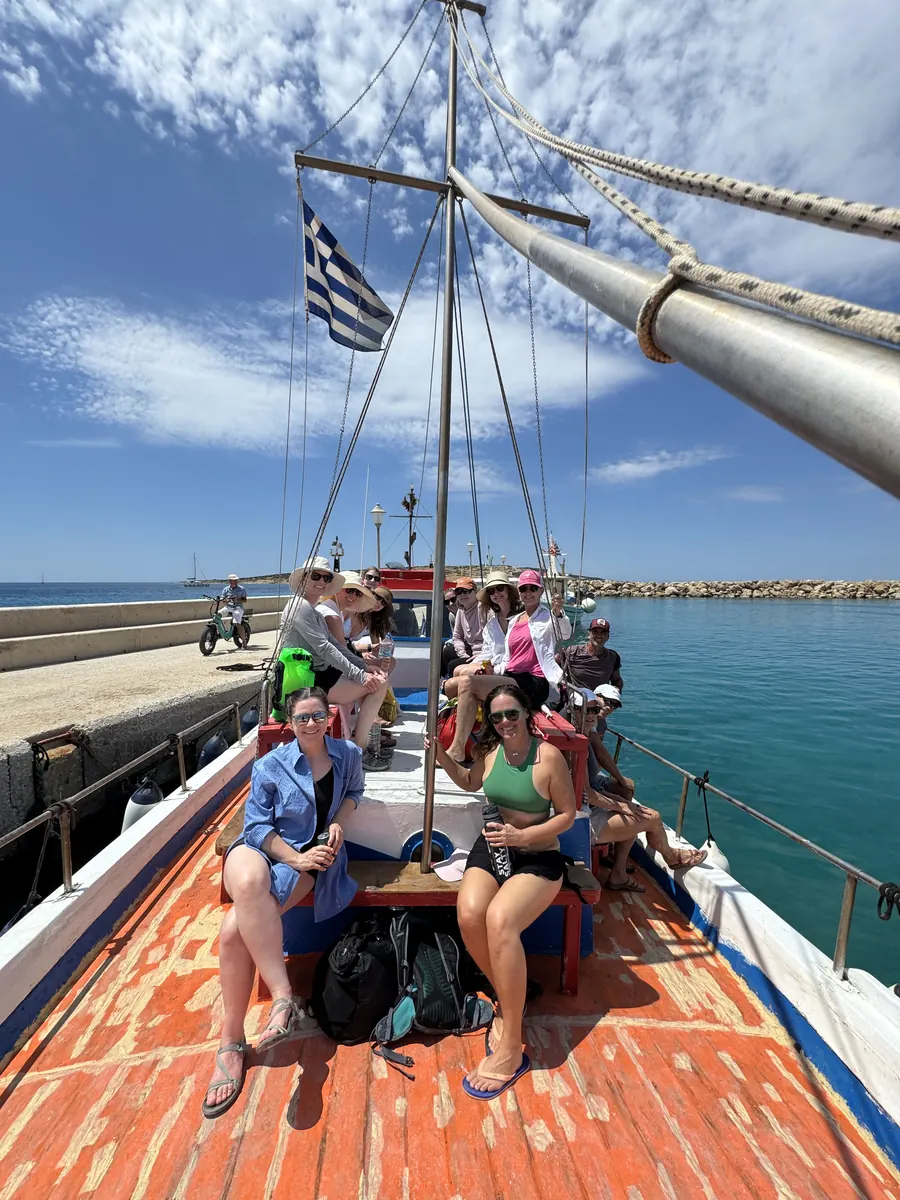 The group seated along the deck of a sailing boat under a Greek flag