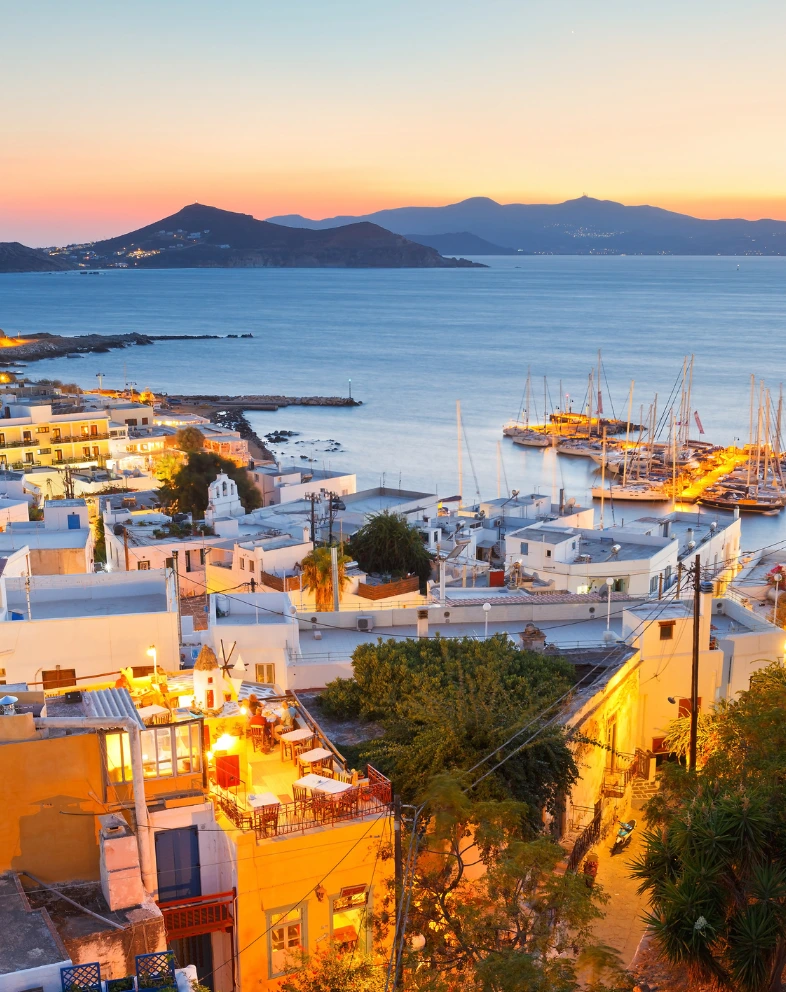 Sunset view over Naxos with whitewashed buildings, harbor, and mountains in the distance.