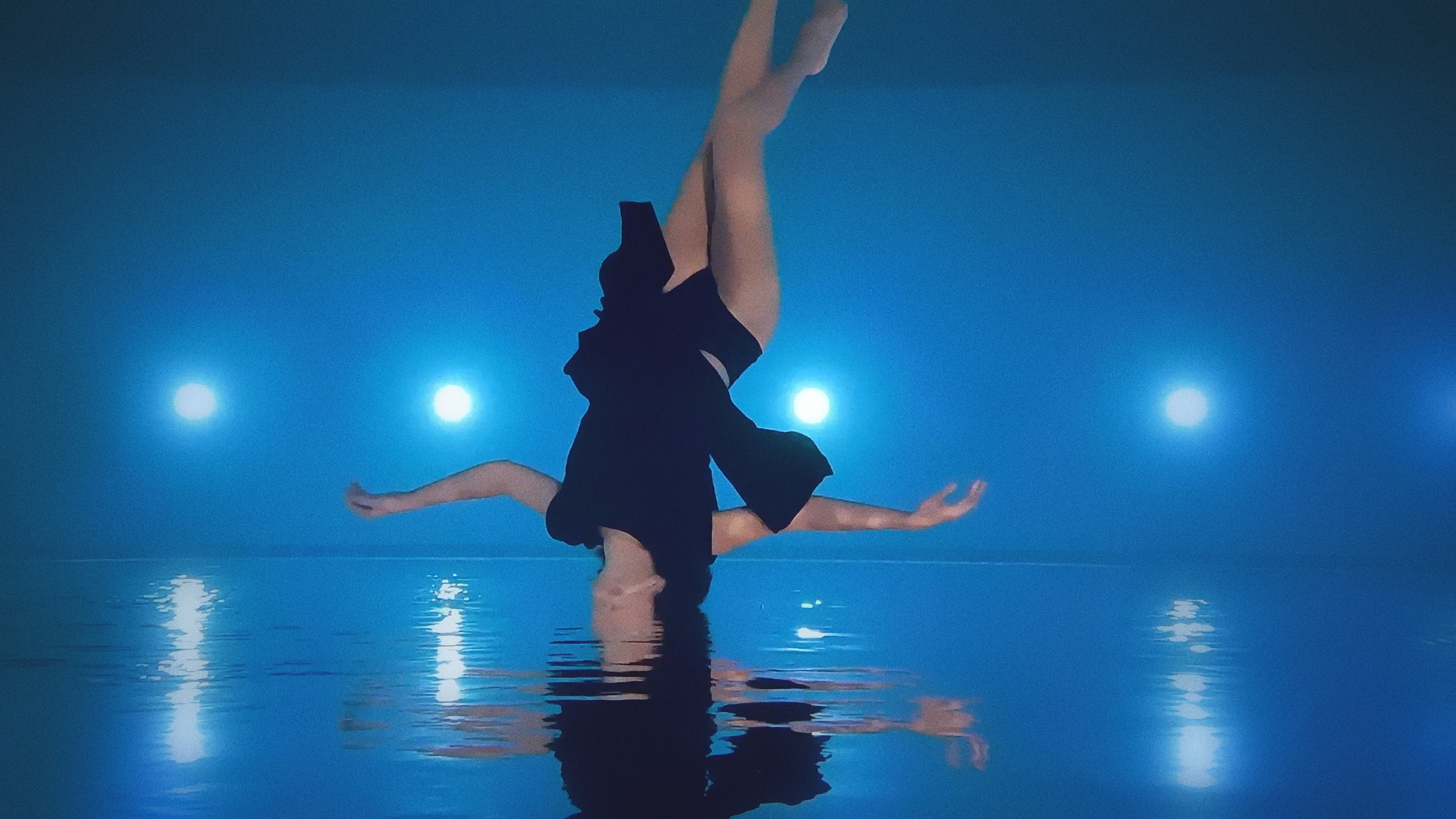 A woman, upside down and wearing a black dress, appears with her head submerged in a pool of water against a blue, spotlit backdrop.