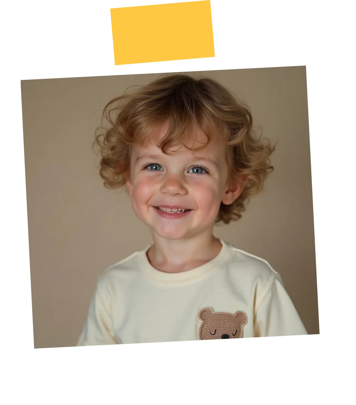 Smiling toddler with curly blonde hair wearing a cream shirt with a teddy bear patch.