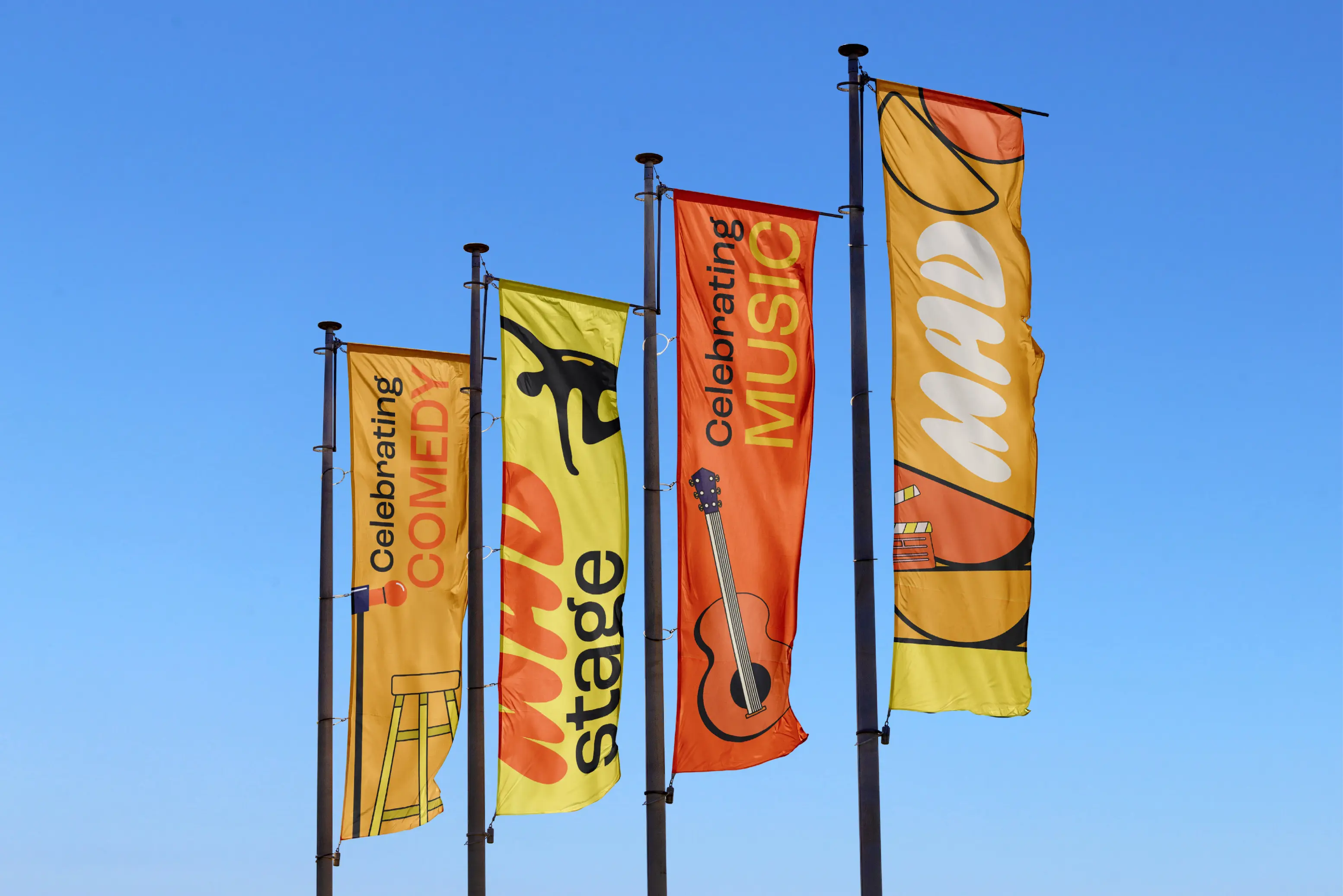 Four vertical flags against a clear blue sky, featuring Celebrating Comedy with a microphone and stool, Mad Stage with a dancing figure, Celebrating Music with a guitar, and Mad with popcorn and a film clapperboard.