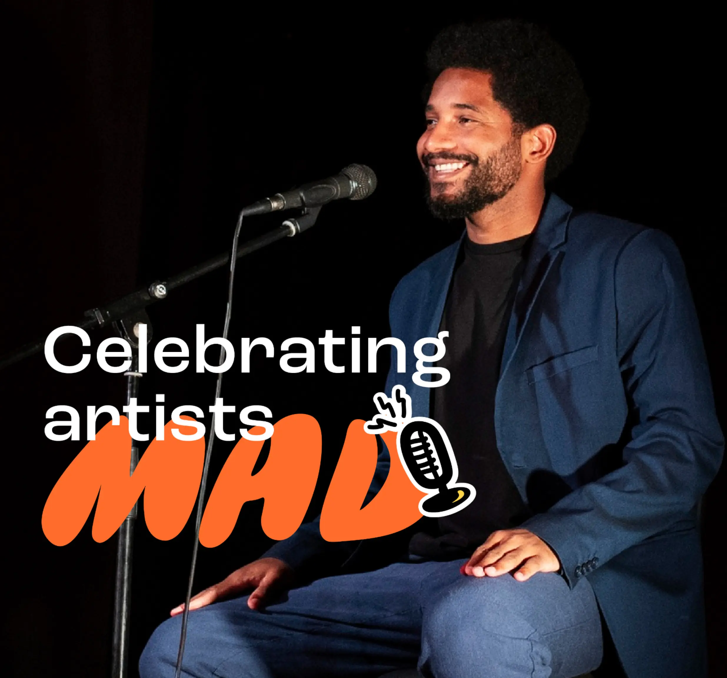 Smiling man in blue suit seated on stage near microphone with text 'Celebrating artists MAD.'