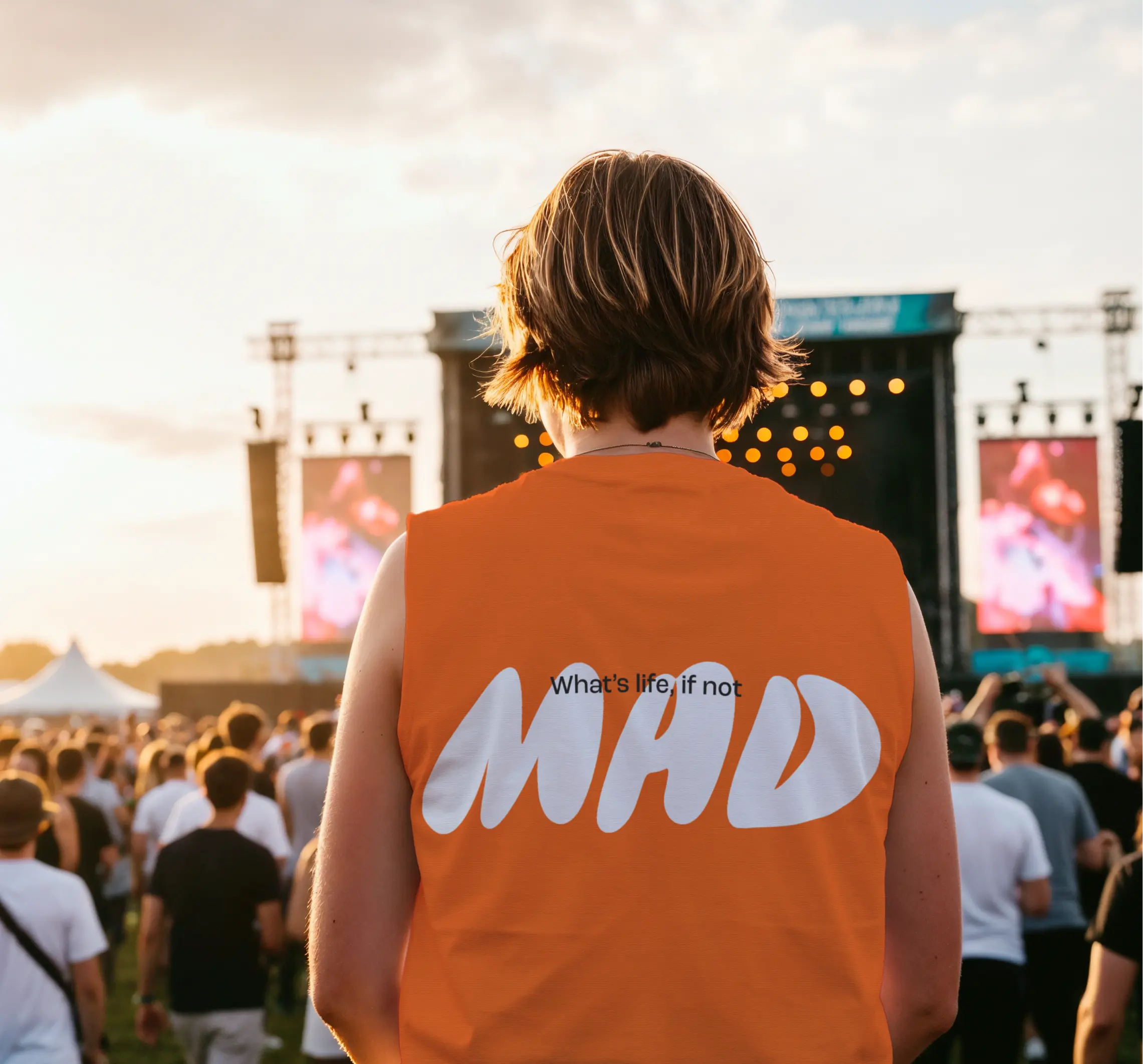 Person with short hair wearing an orange sleeveless shirt printed with 'What's life if not MAD' at an outdoor concert crowd during sunset.