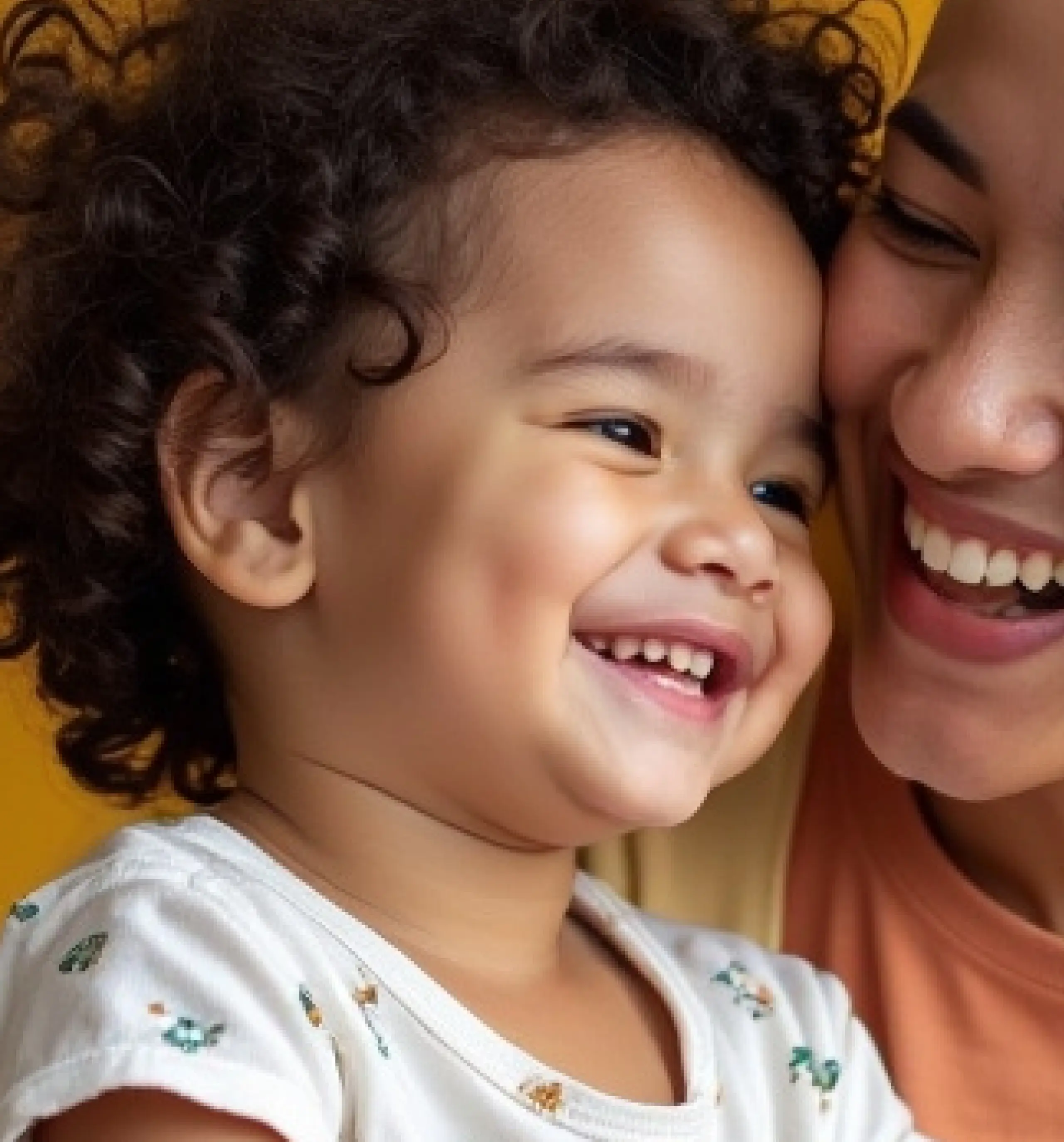 Close-up of a smiling toddler with curly hair leaning against a woman who is also smiling.