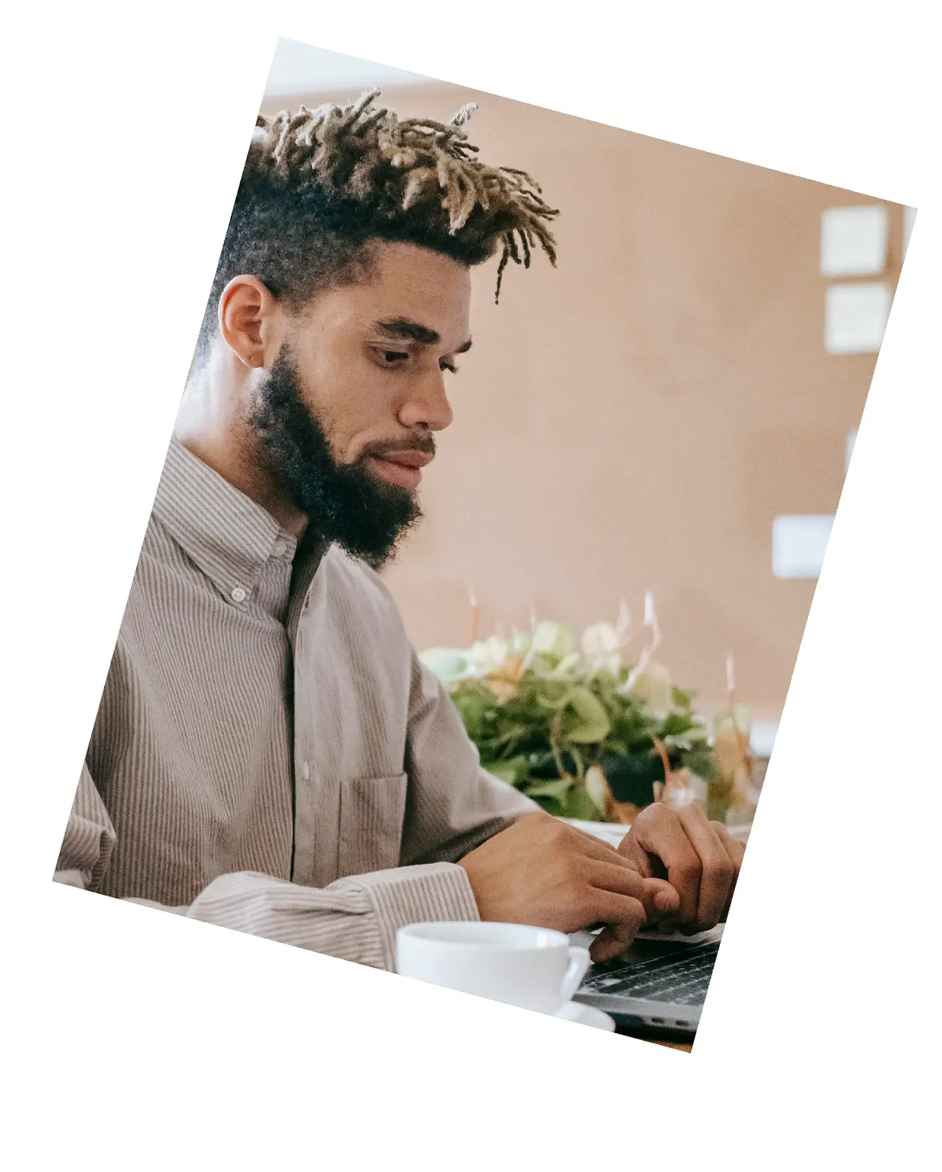 Man with a beard and dreadlocks wearing a striped shirt working on a laptop at a table with a cup and plants in the background.