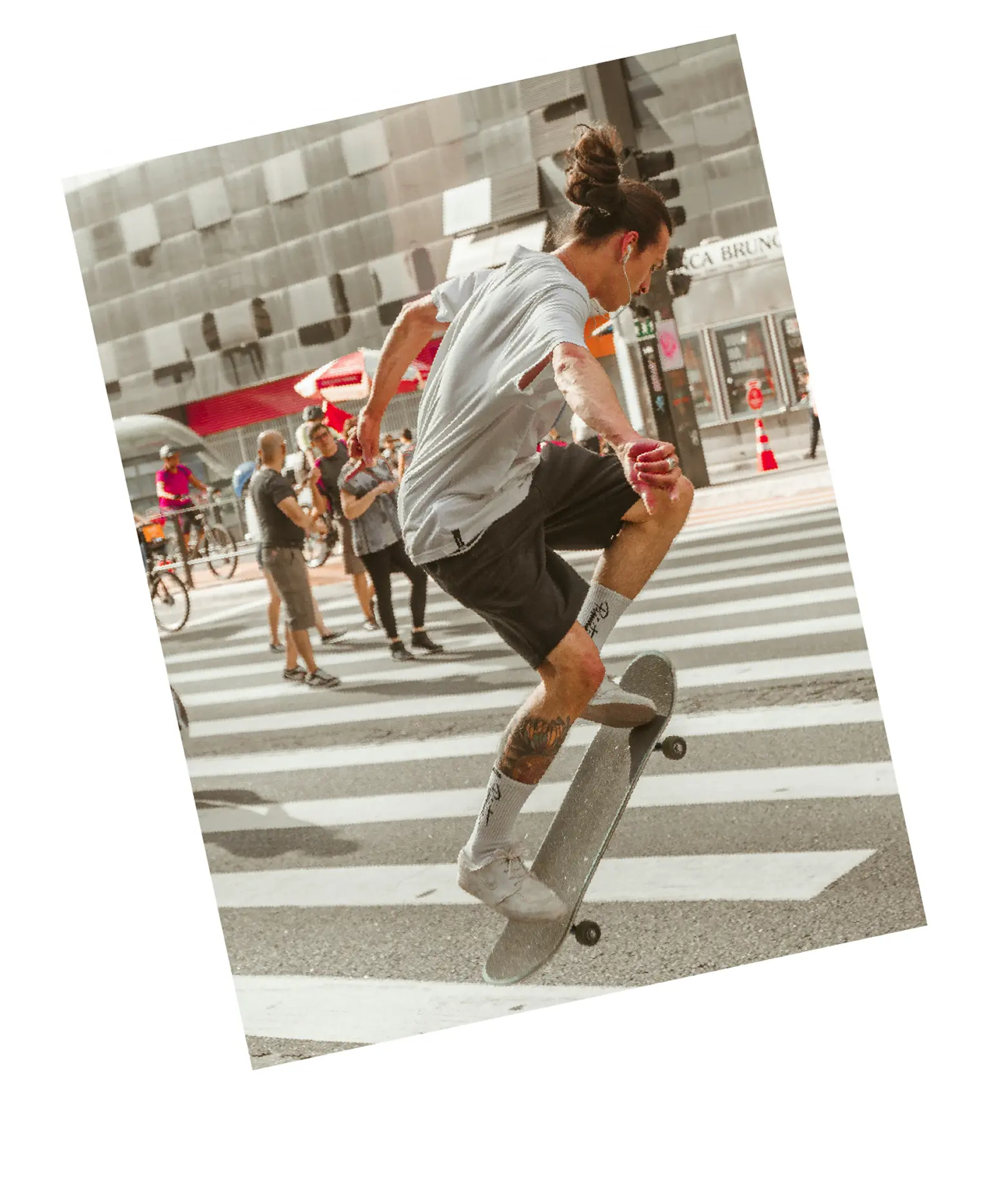Young man with a bun and tattoos performing a skateboard trick on a city crosswalk with people and cyclists in the background.