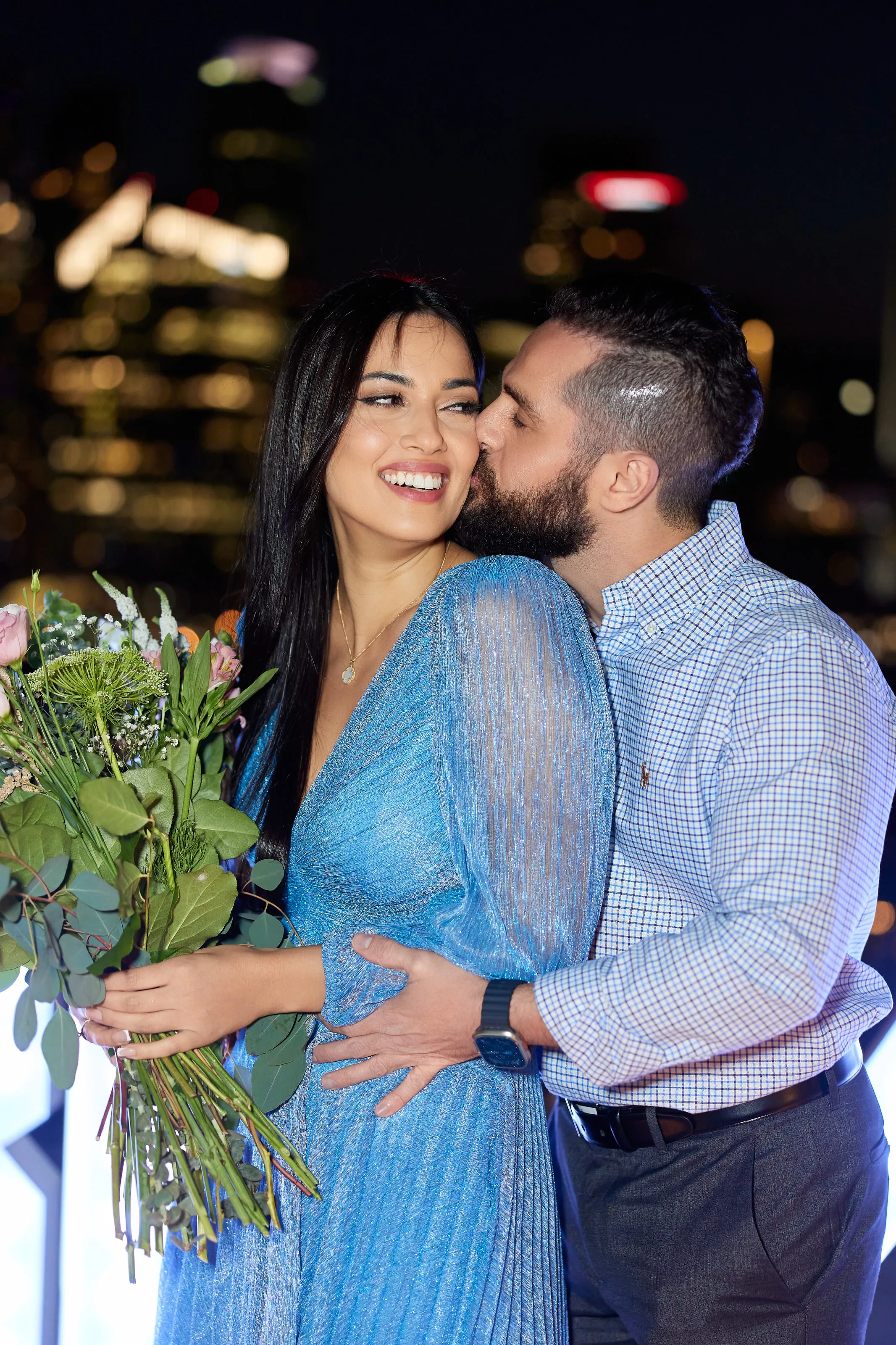 Un homme embrasse la joue de sa fiancée souriante, fleurs à la main, devant les lumières de la ville la nuit
