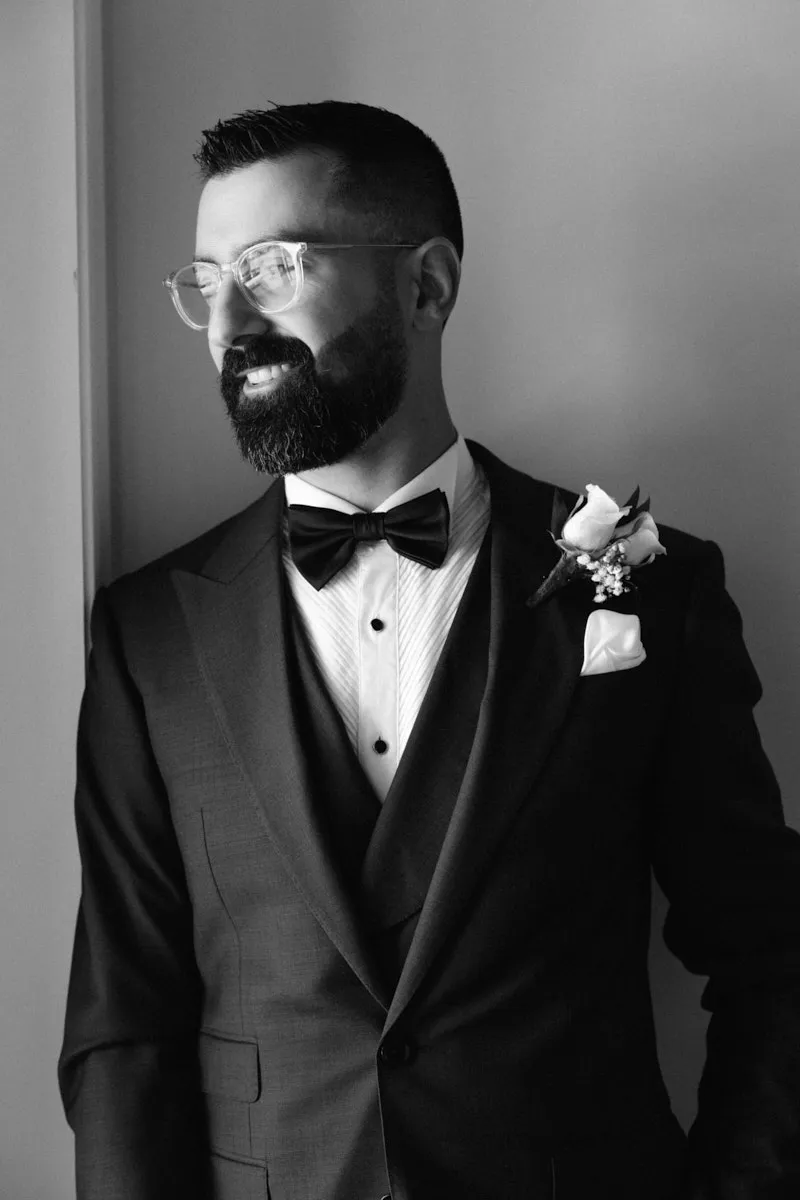 Portrait en noir et blanc d'un marié souriant avec lunettes, nœud papillon et boutonnière près d'une fenêtre
