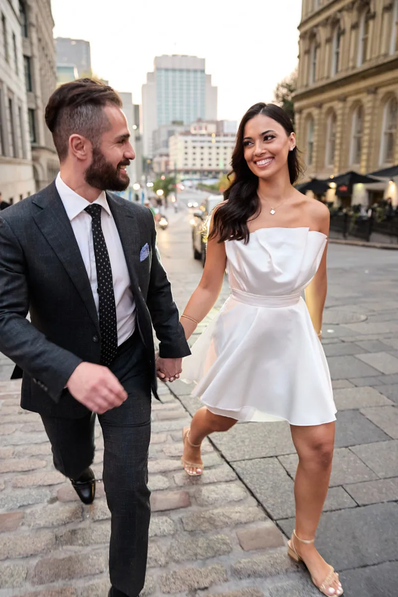 Couple, elle en robe blanche courte et lui en complet gris, marchant main dans la main dans une rue pavée du Vieux-Montréal