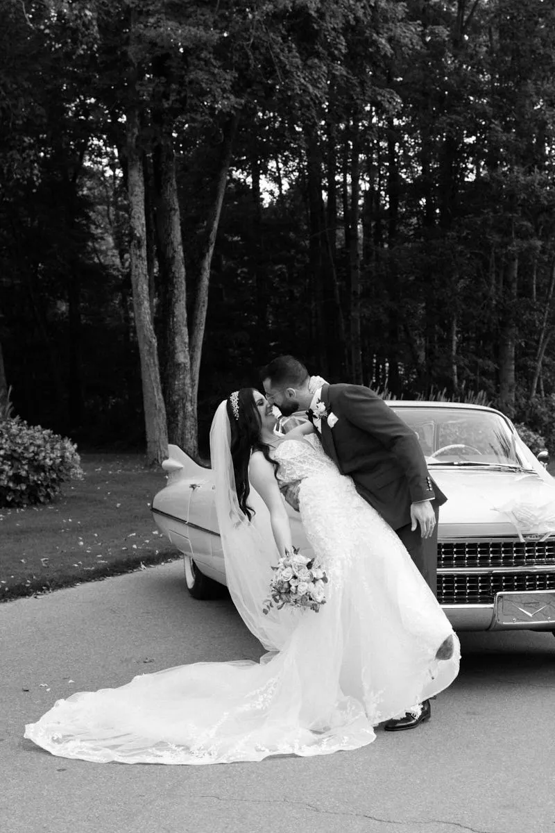 Photo en noir et blanc du marié embrassant la mariée près d'une voiture ancienne, son long voile au sol