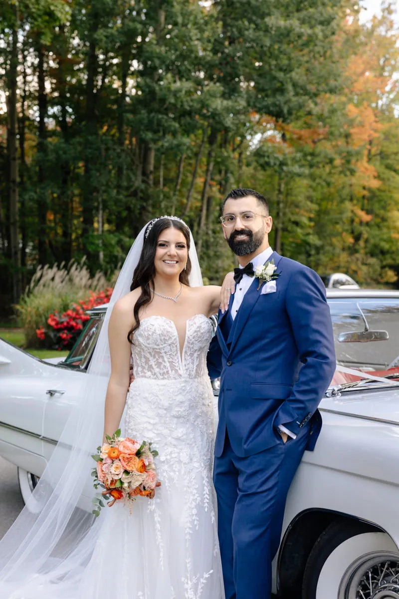 La mariée et le marié en complet bleu posant près d'une voiture ancienne blanche sous des arbres d'automne
