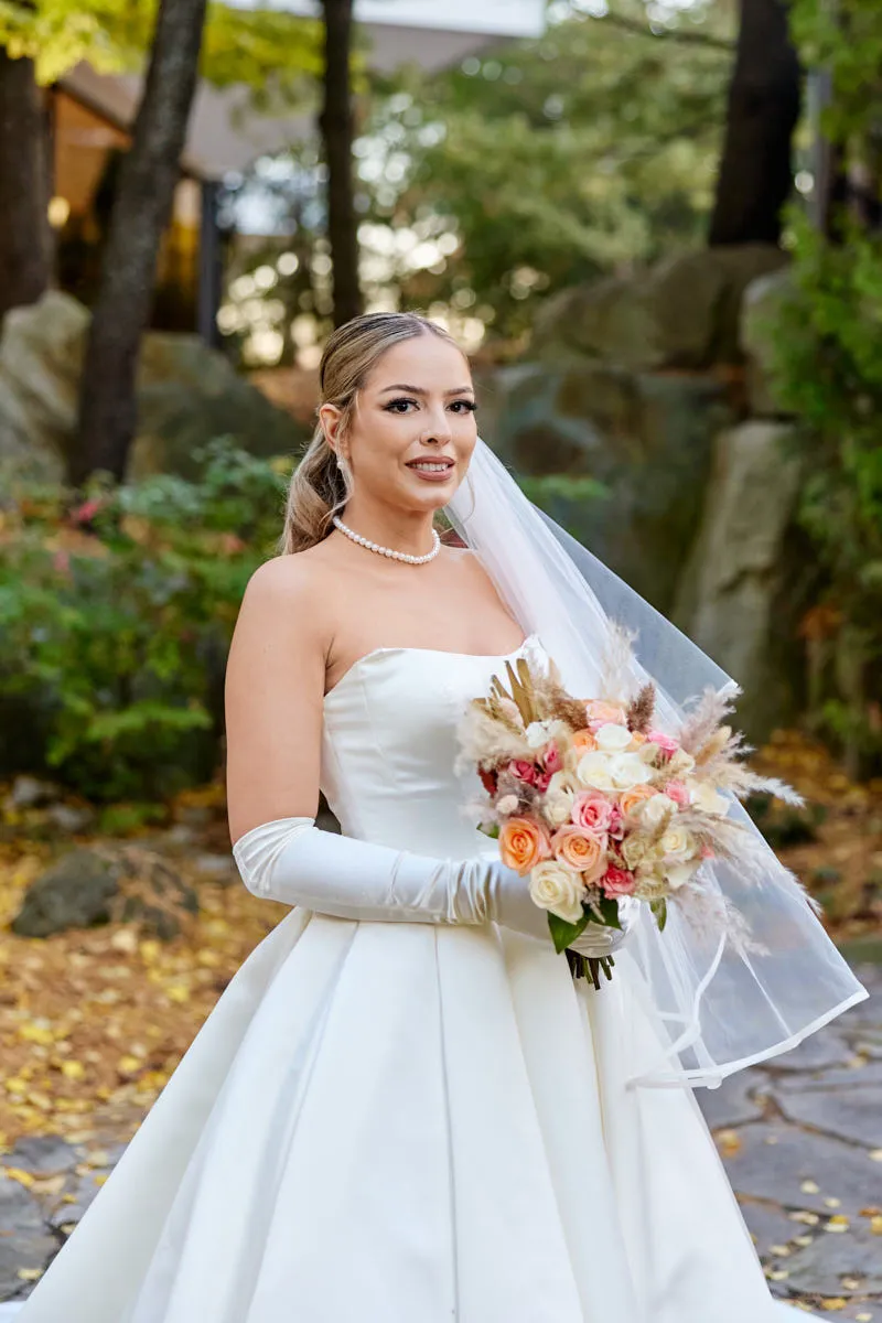 Mariée en robe bustier de satin, longs gants et perles, tenant un bouquet pêche et rose
