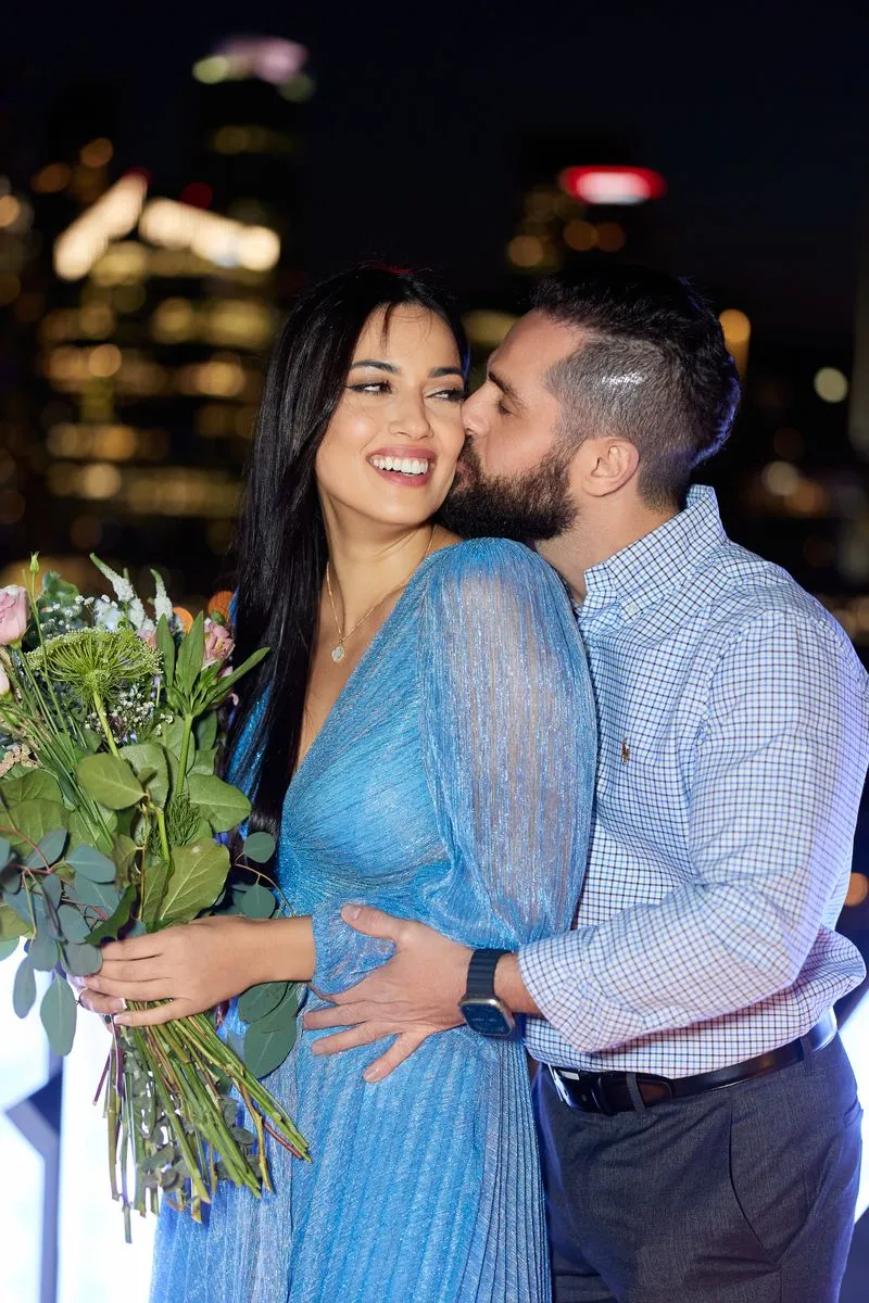 Man kissing the cheek of his smiling fiancée holding flowers, city lights at night behind them