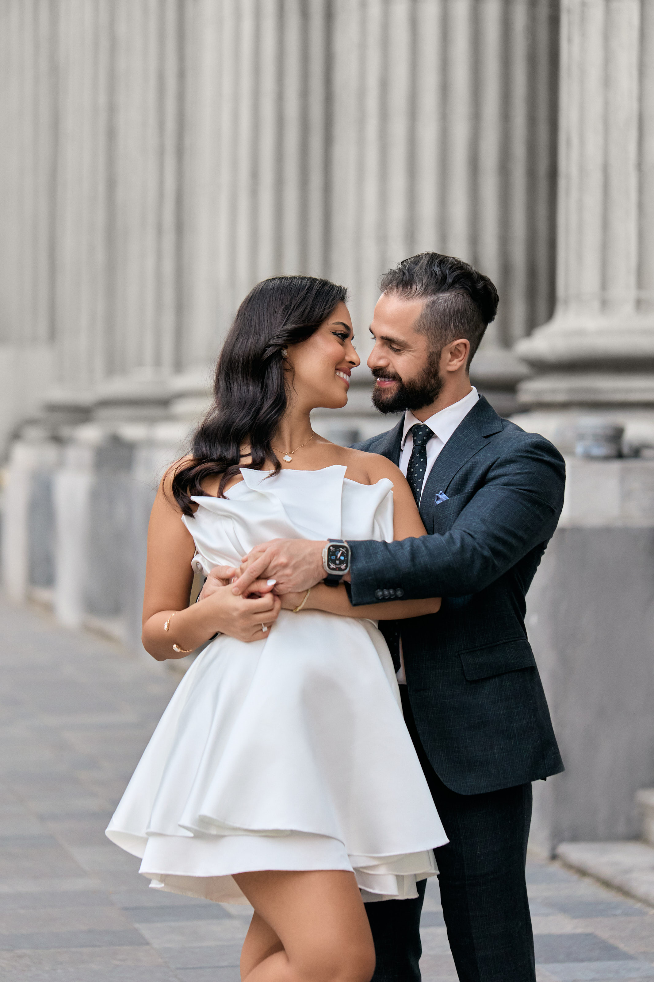 Couple in a short white dress and navy suit embracing in front of stone columns in Old Montreal