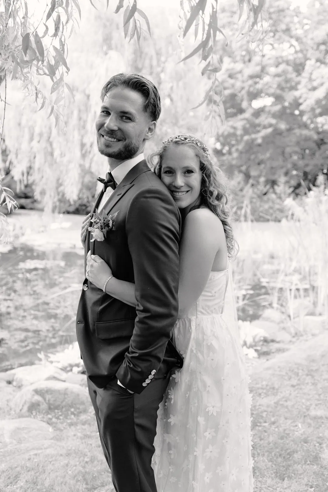 Black-and-white photo of a bride hugging her groom from behind beneath willow trees