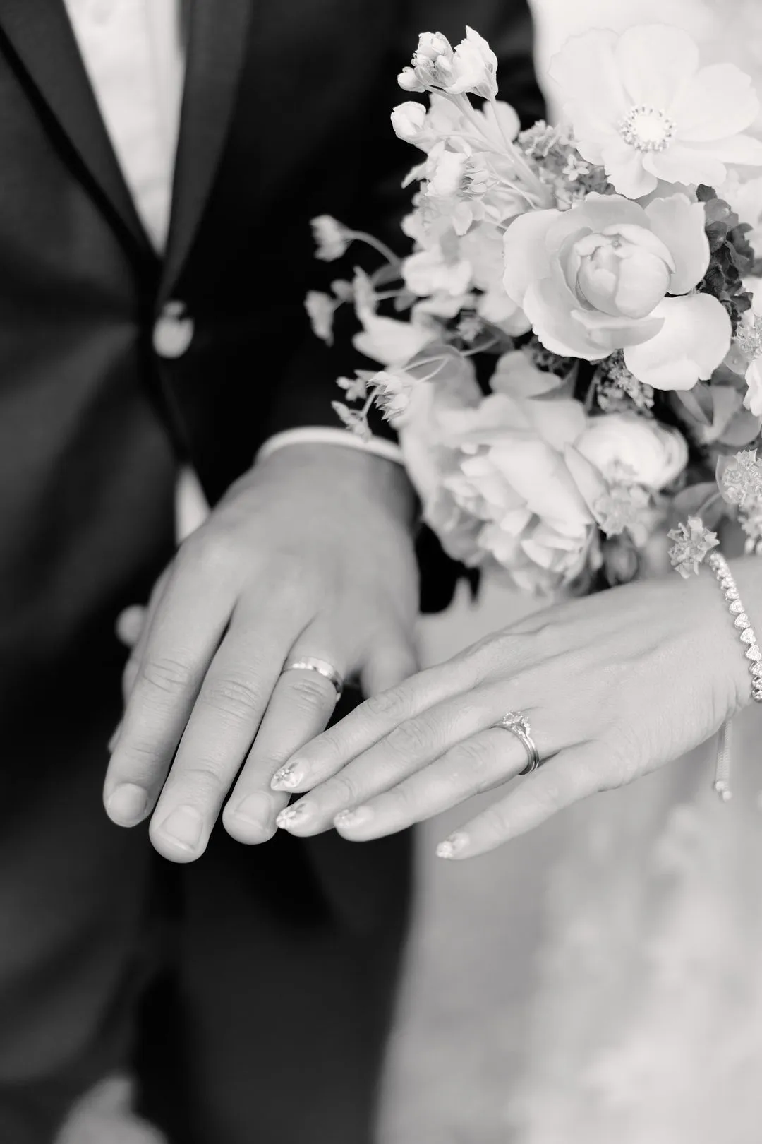 Black-and-white close-up of the couple's hands showing their wedding rings and the bouquet