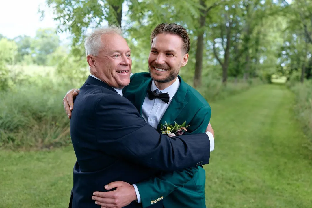 Groom in a green tuxedo sharing a big hug with an older man outdoors