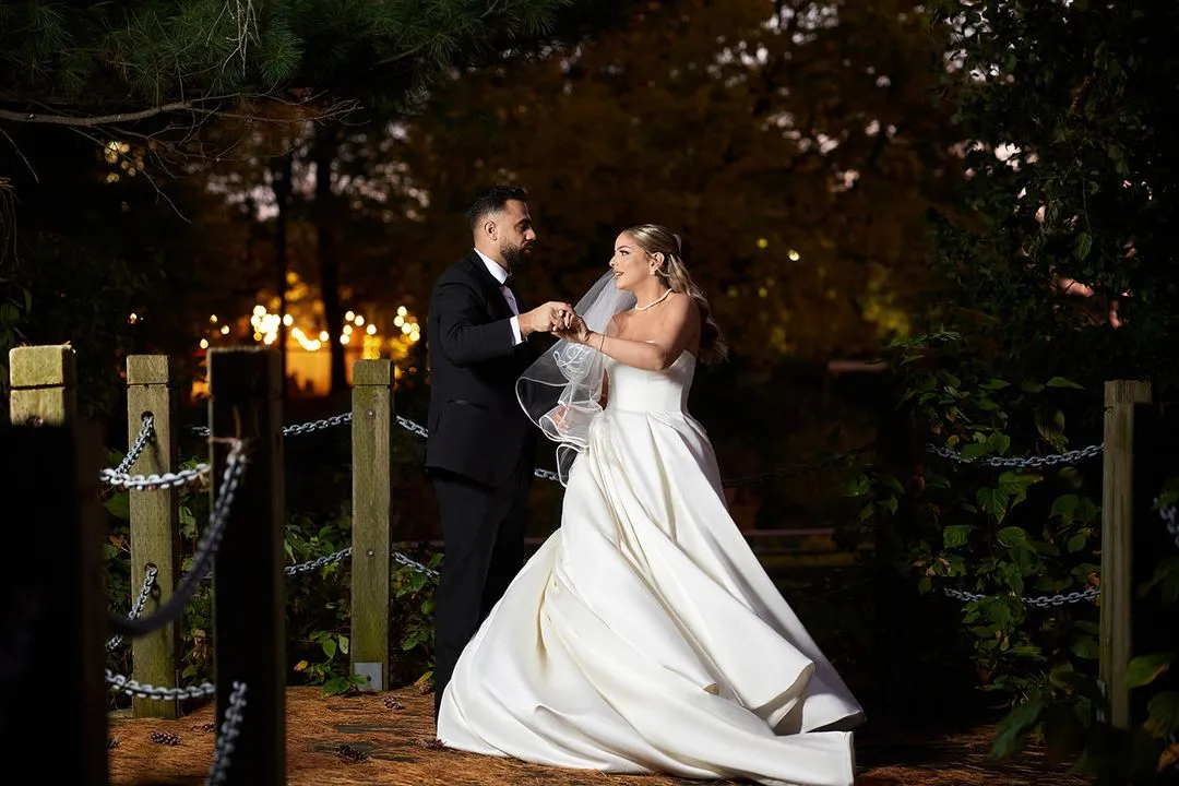 Groom adjusting the bride's veil at dusk, warm lights glowing in the trees behind them