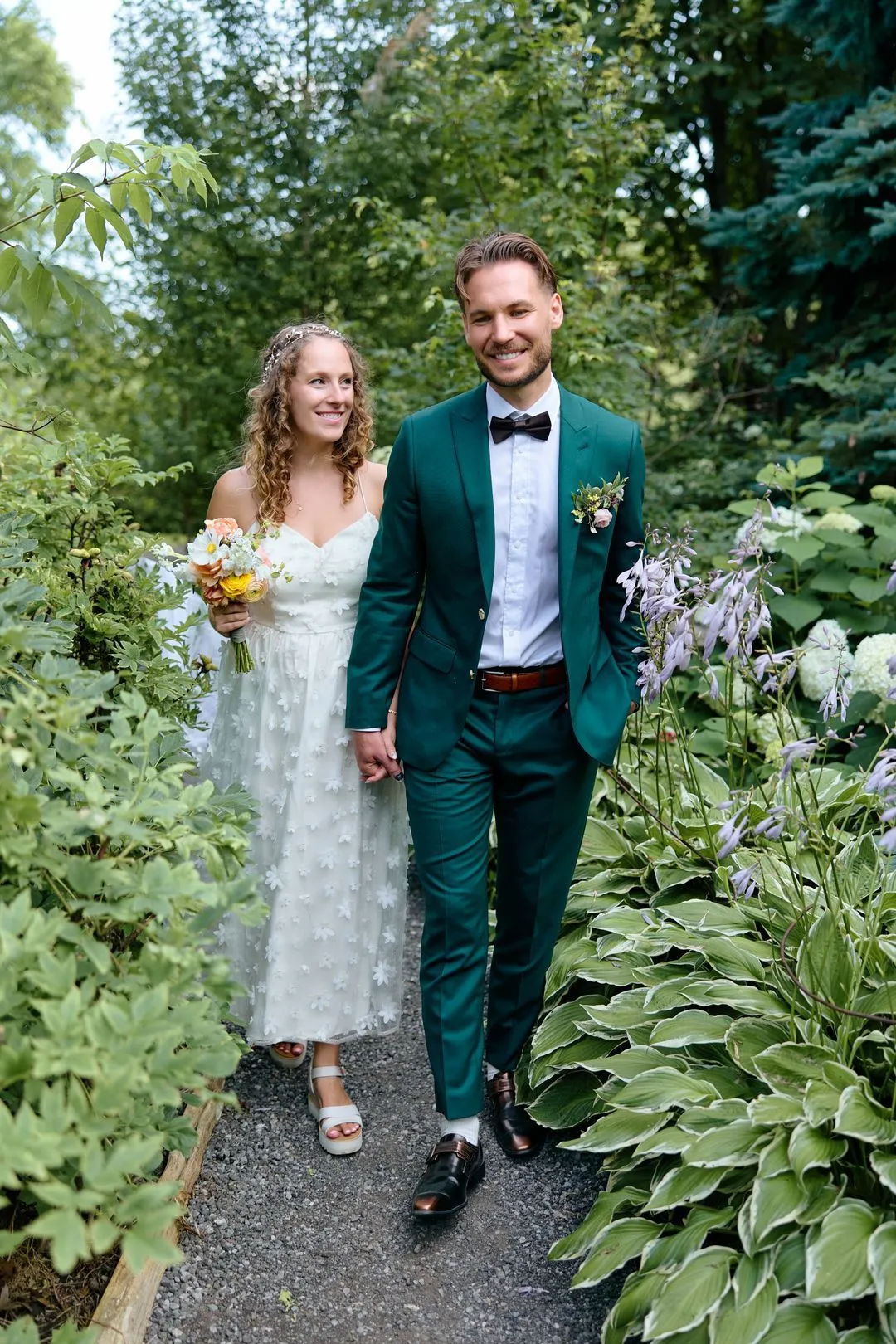 Groom in a green suit walking hand in hand with the bride along a leafy garden path