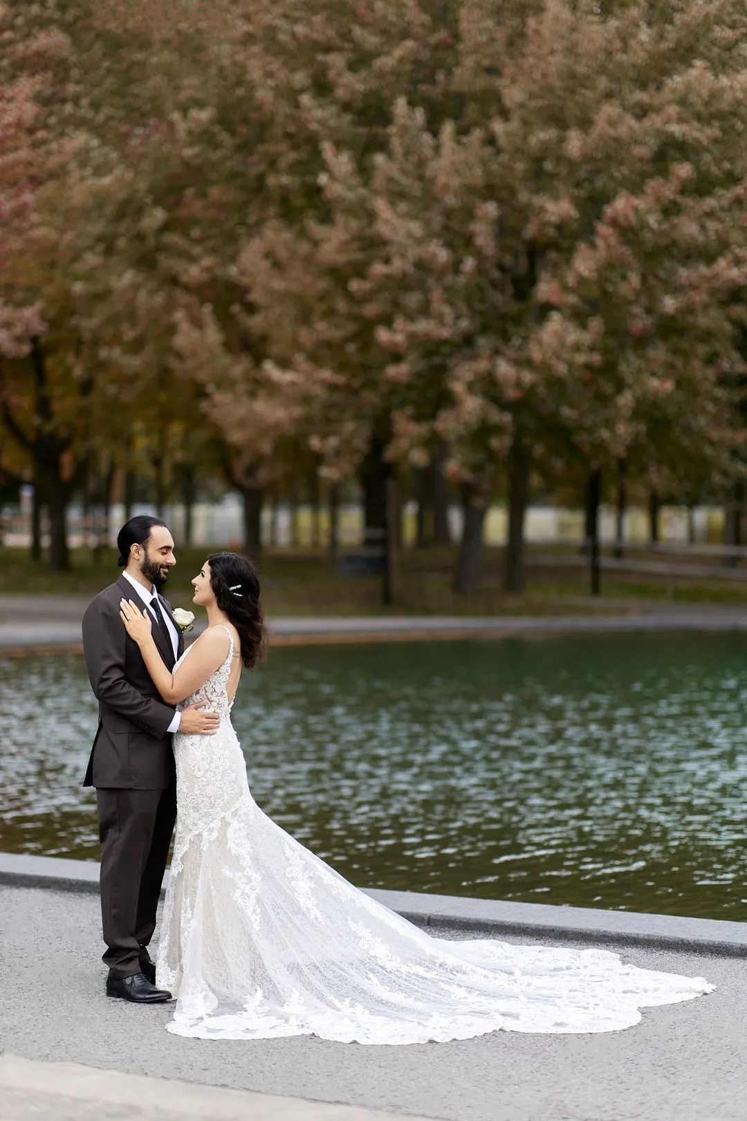 Bride and groom embracing by a reflecting pool lined with autumn trees
