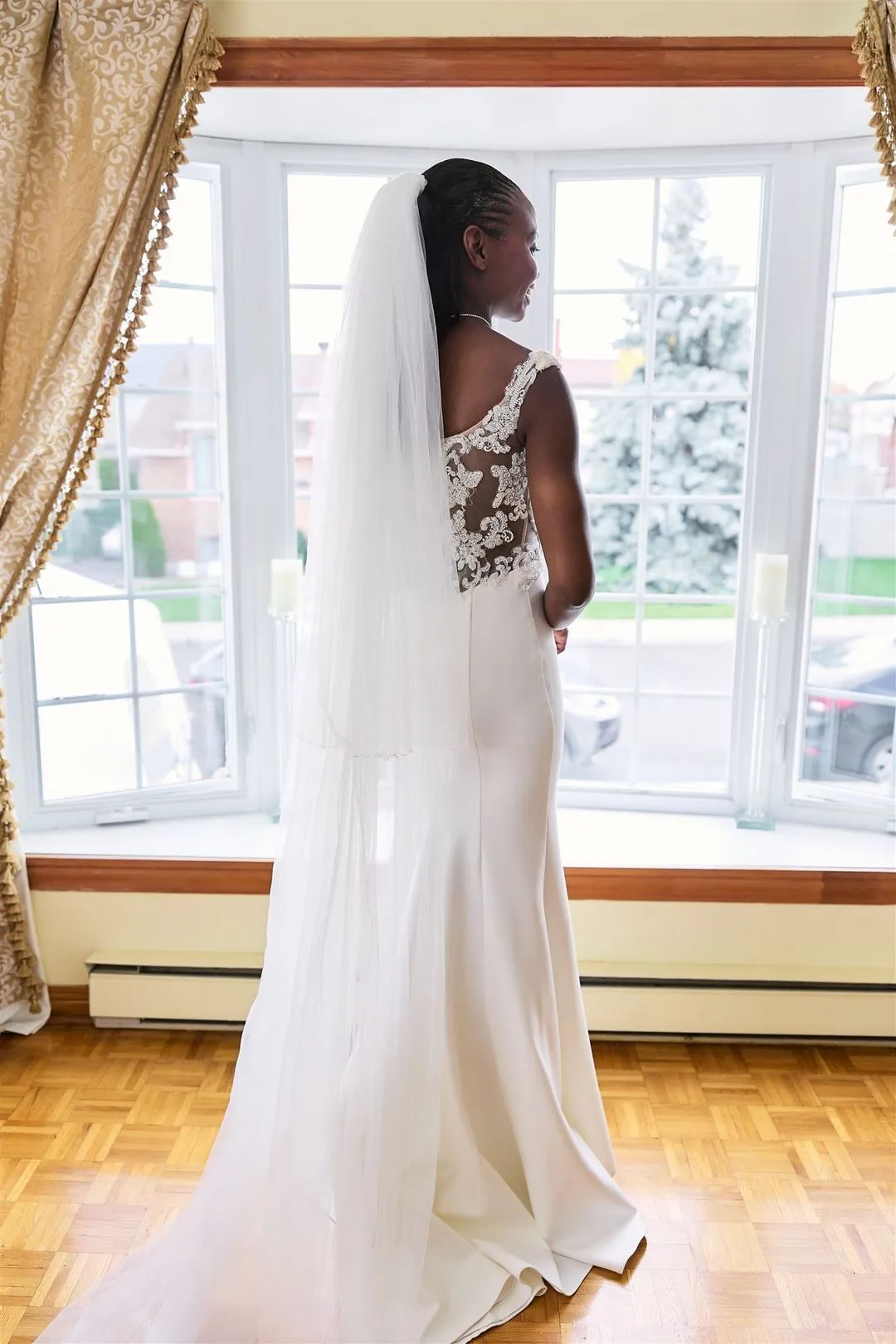 Bride in her gown and long veil looking out a bay window while getting ready