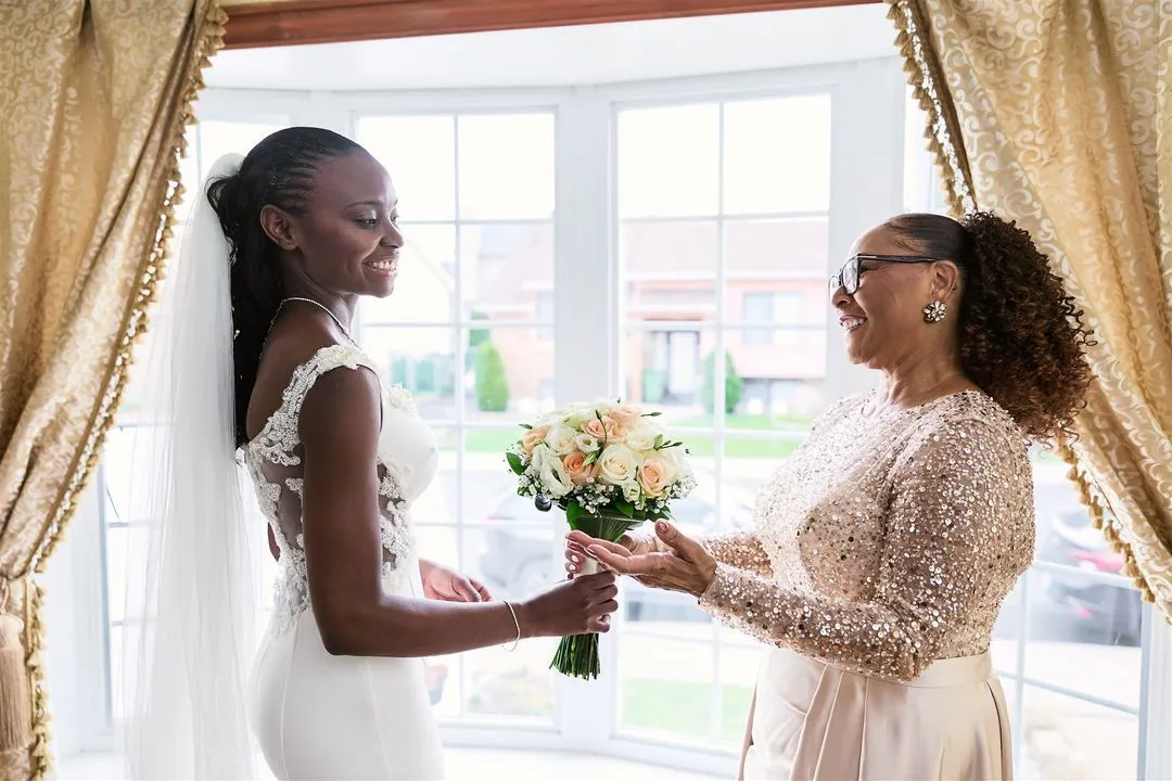 Bride receiving her bouquet from a smiling woman by the window while getting ready