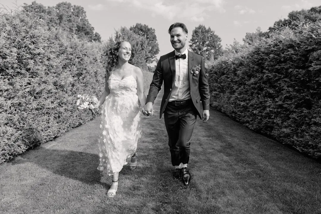 Black-and-white photo of a bride and groom walking hand in hand between tall hedges