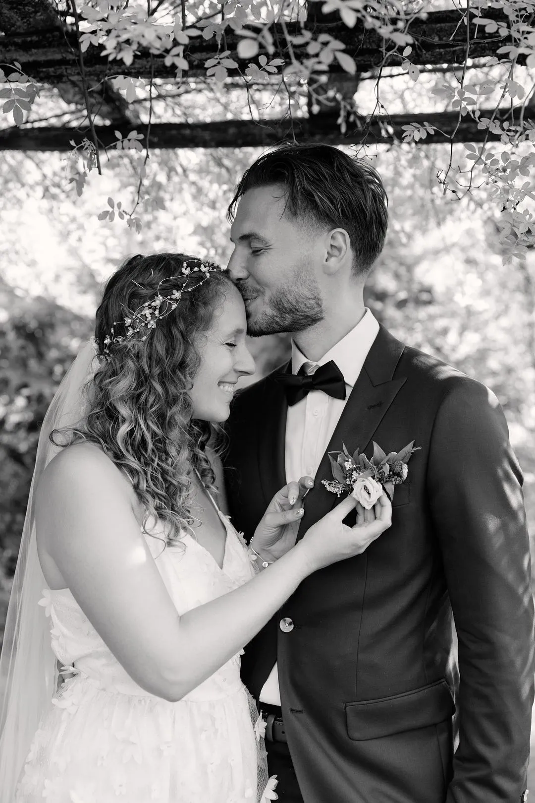 Black-and-white photo of the groom kissing the bride's forehead under a garden pergola