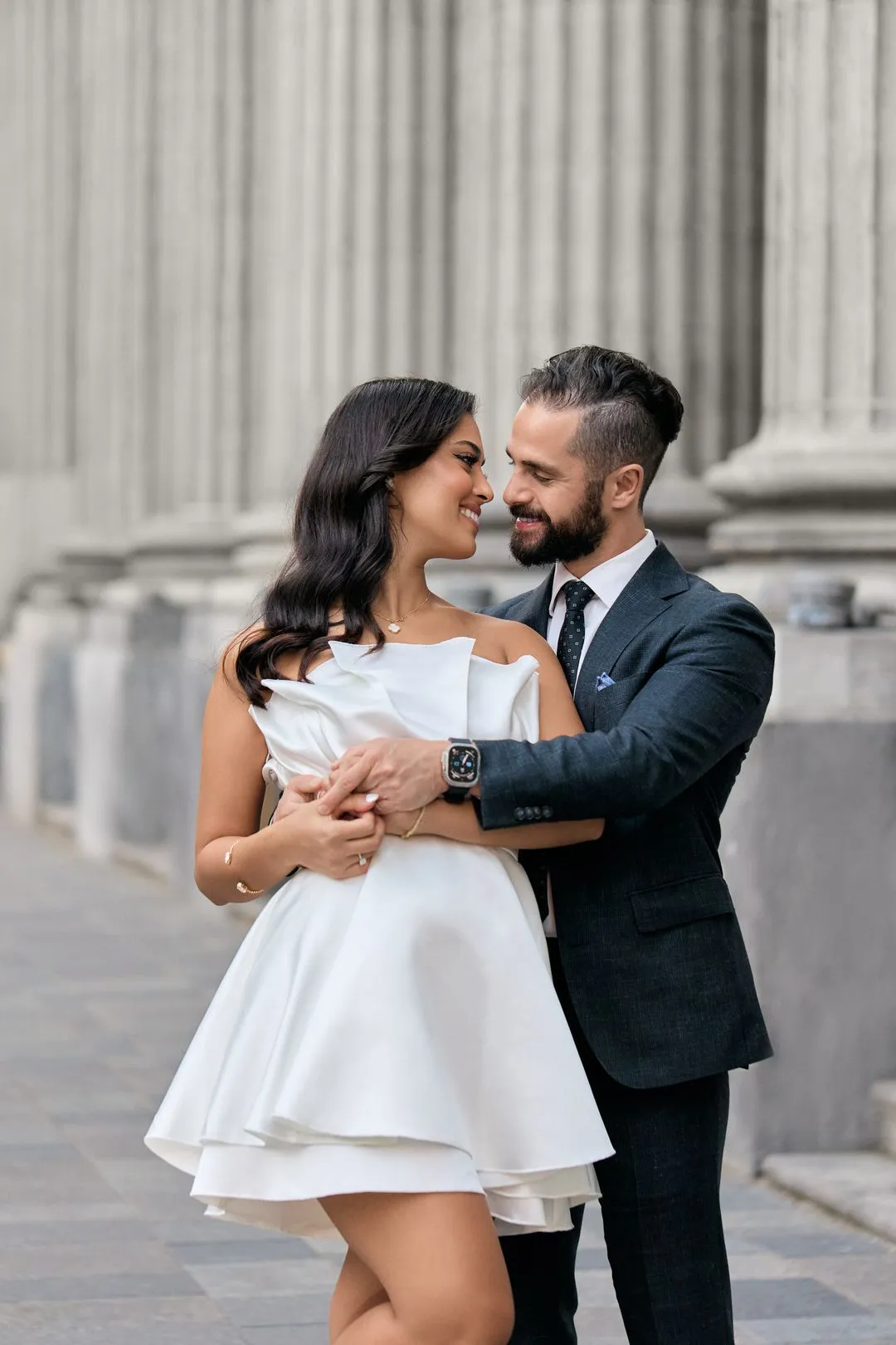 Couple, elle en robe blanche courte et lui en complet marine, enlacé devant des colonnes de pierre dans le Vieux-Montréal