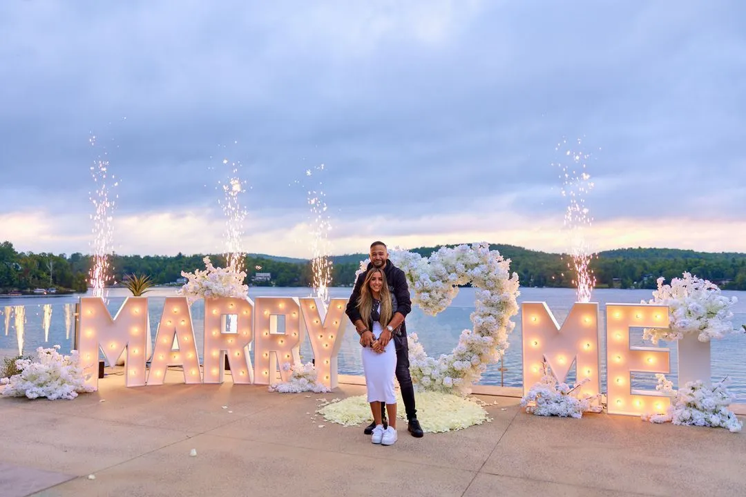 Couple enlacé devant une enseigne lumineuse MARRY ME et des feux de Bengale au bord d'un lac