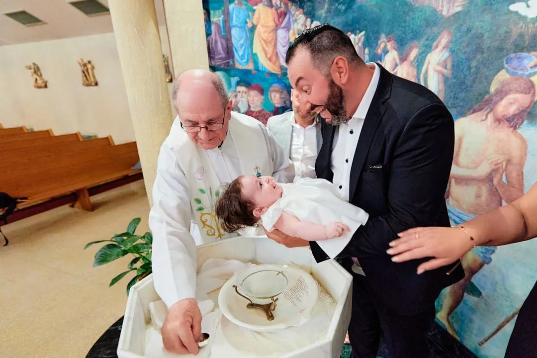 Un prêtre célèbre un baptême aux fonts baptismaux pendant qu'un homme tient le bébé vêtu de blanc