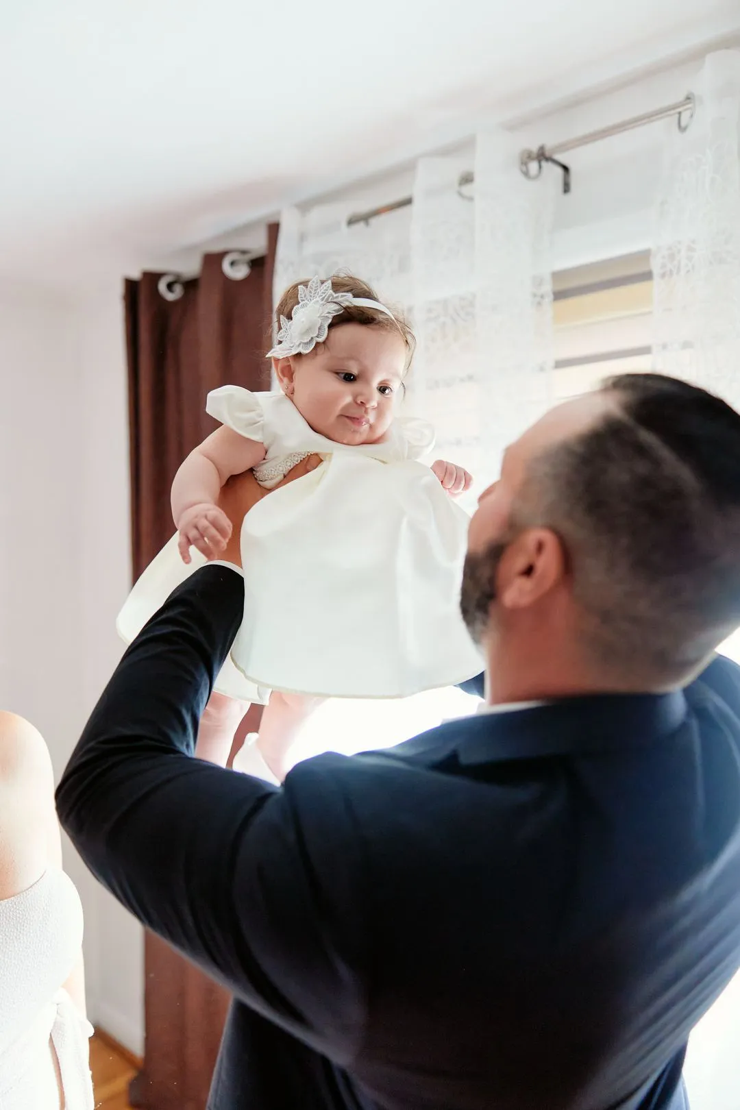 Un homme soulève un bébé souriant en robe de baptême blanche près de la fenêtre