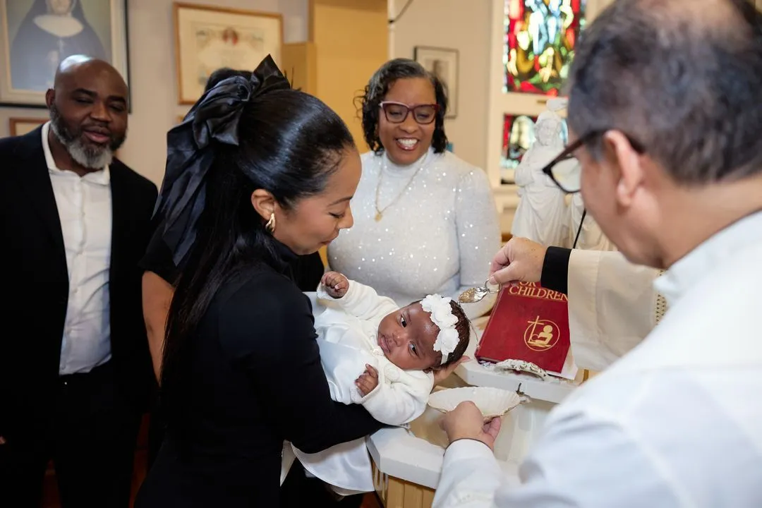 Le prêtre baptise un bébé coiffé d'un bonnet blanc, tenu au-dessus des fonts par sa famille
