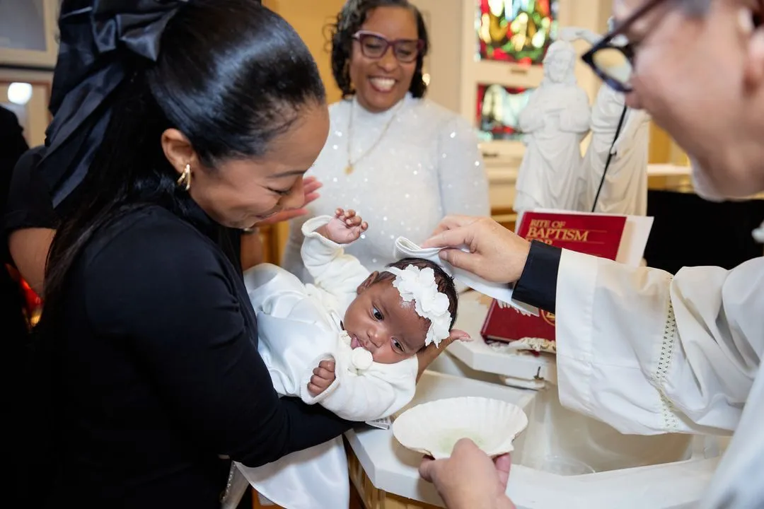 Bébé avec un nœud blanc levant les yeux pendant que le prêtre verse l'eau du baptême