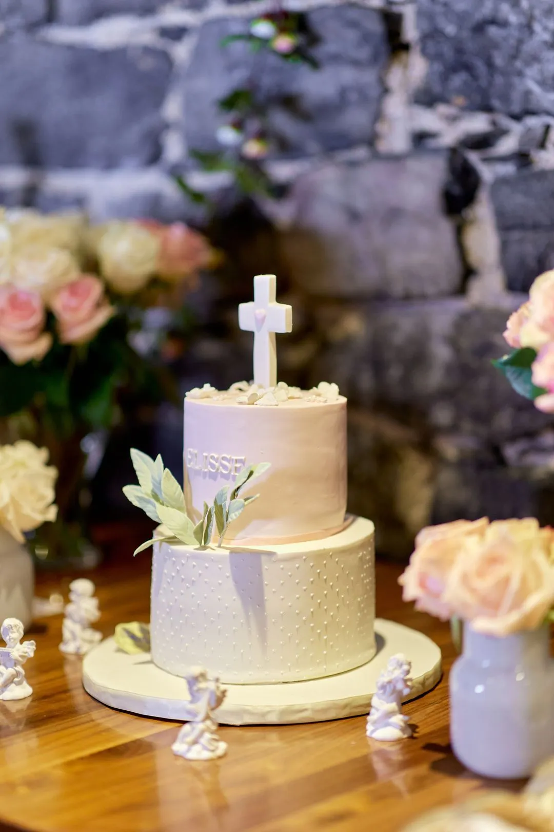 Gâteau de baptême blanc à deux étages orné d'une croix, entouré de roses et de figurines d'anges