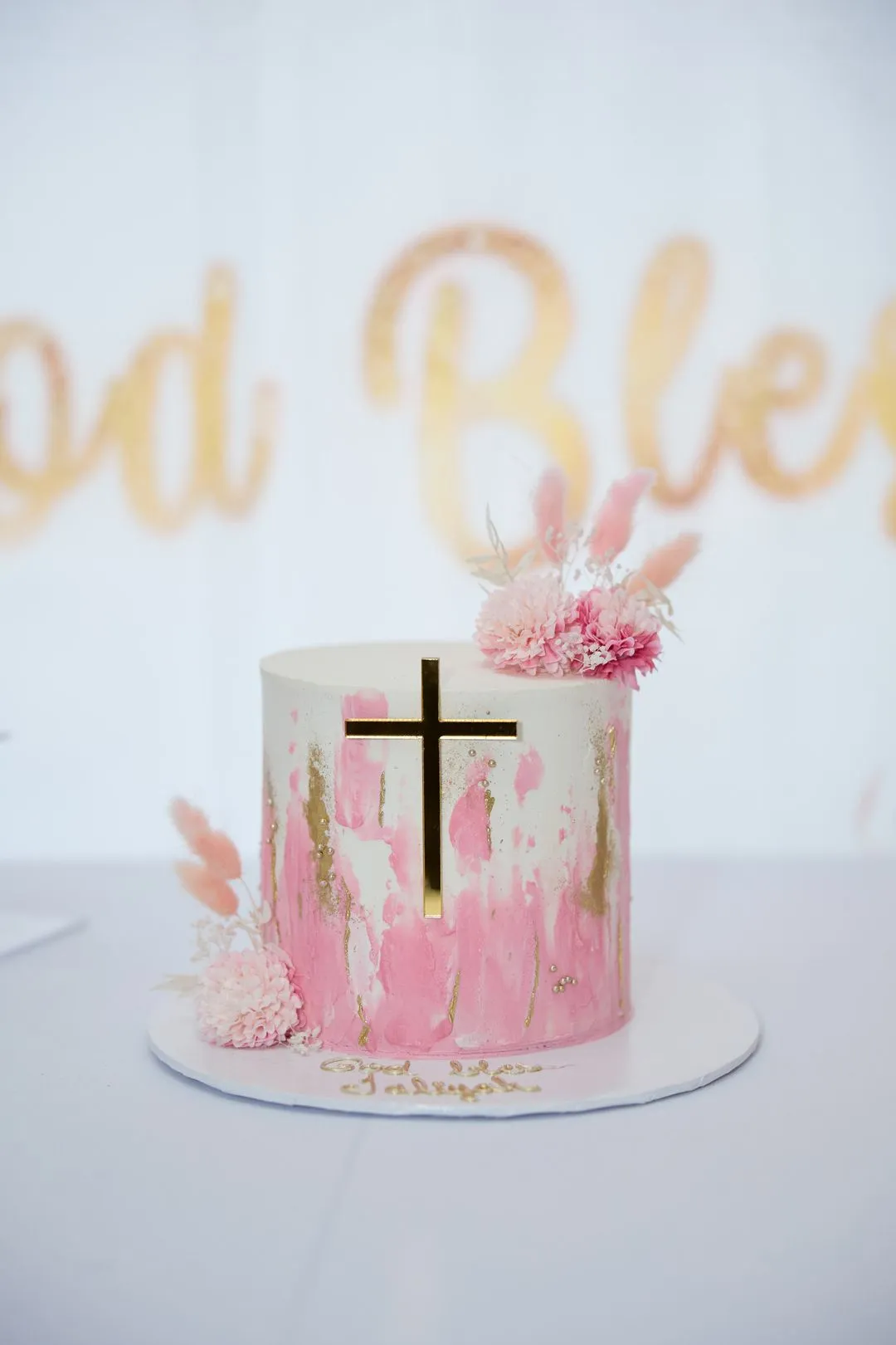 Gâteau de baptême rose et blanc avec une croix dorée devant une enseigne God Bless