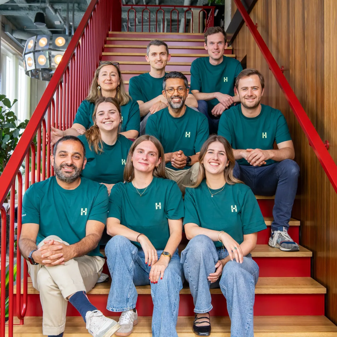 Group of nine smiling people wearing matching green shirts sitting on red stairs indoors.