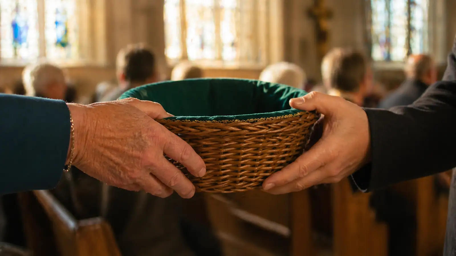 Close-up warm photograph of a woven offering basket being passed between hands in a sunlit French church nave, soft golden light filtering through stained glass windows, shallow depth of field, teal and warm tones, photorealistic, editorial style --ar 16:9 --style raw