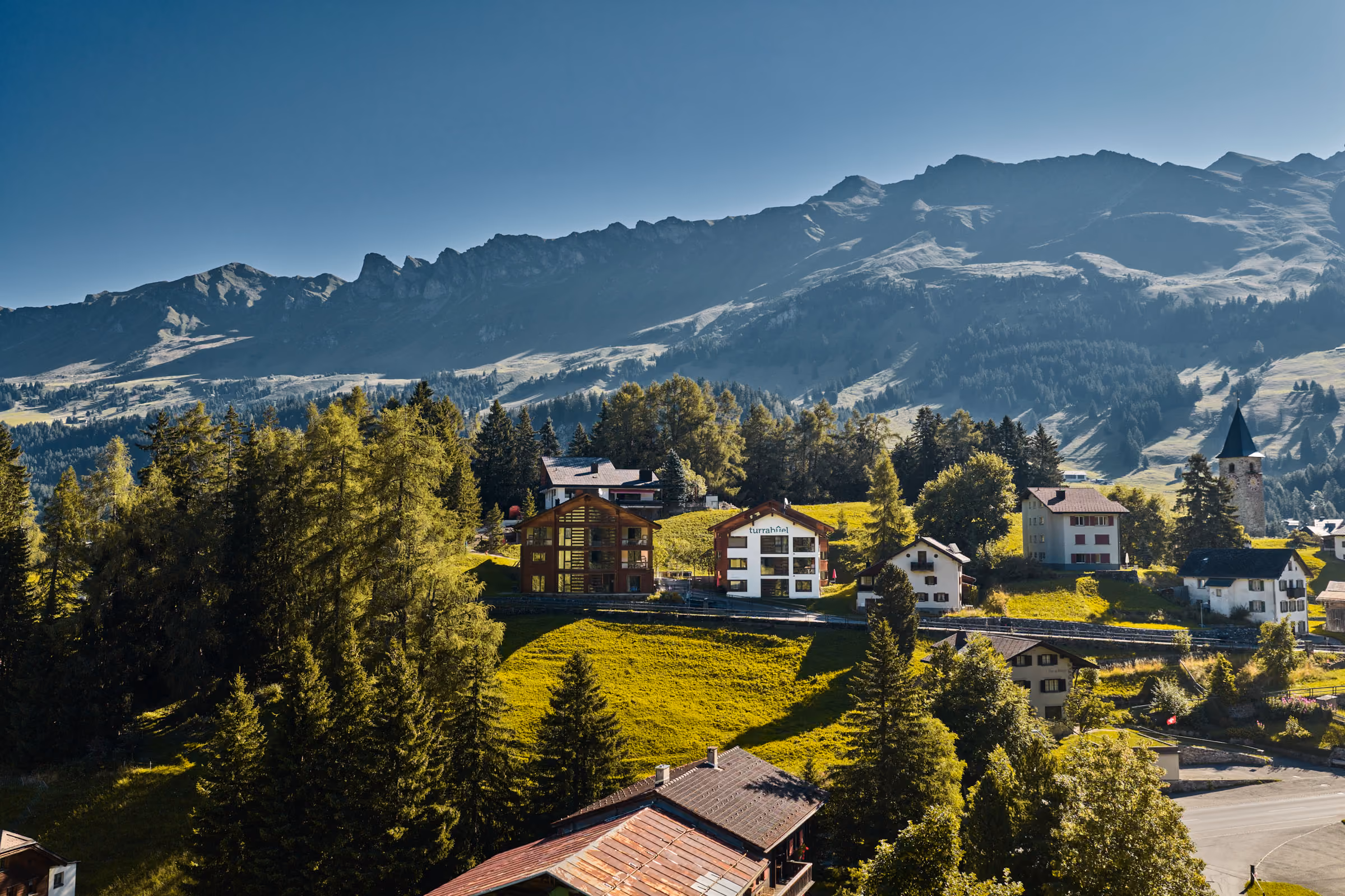 Die zwei Häuser der Turrabuel Ferienwohnungen mitten in saftig-grünen Bergwiesen in der Region Lenzerheide. Im Hintergrund sind Berge der Region wie das Schwarzhorn zu sehen.