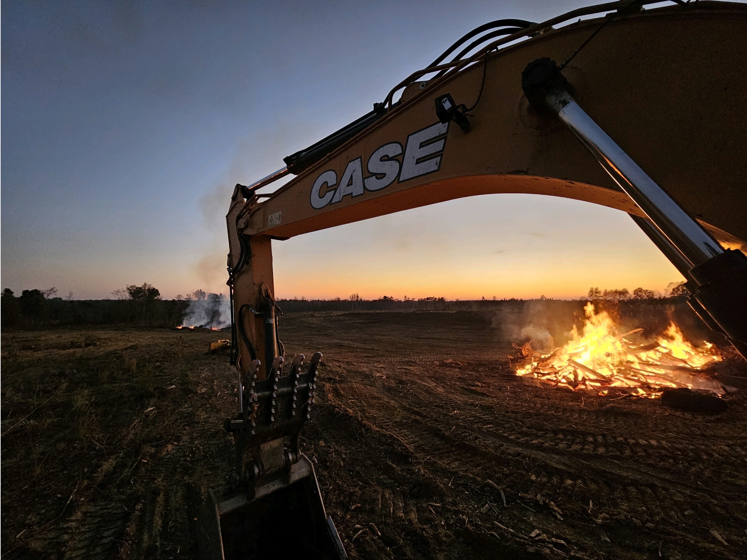 Excavator arm labeled CASE in front of a controlled burn fire on a dirt field at sunset.
