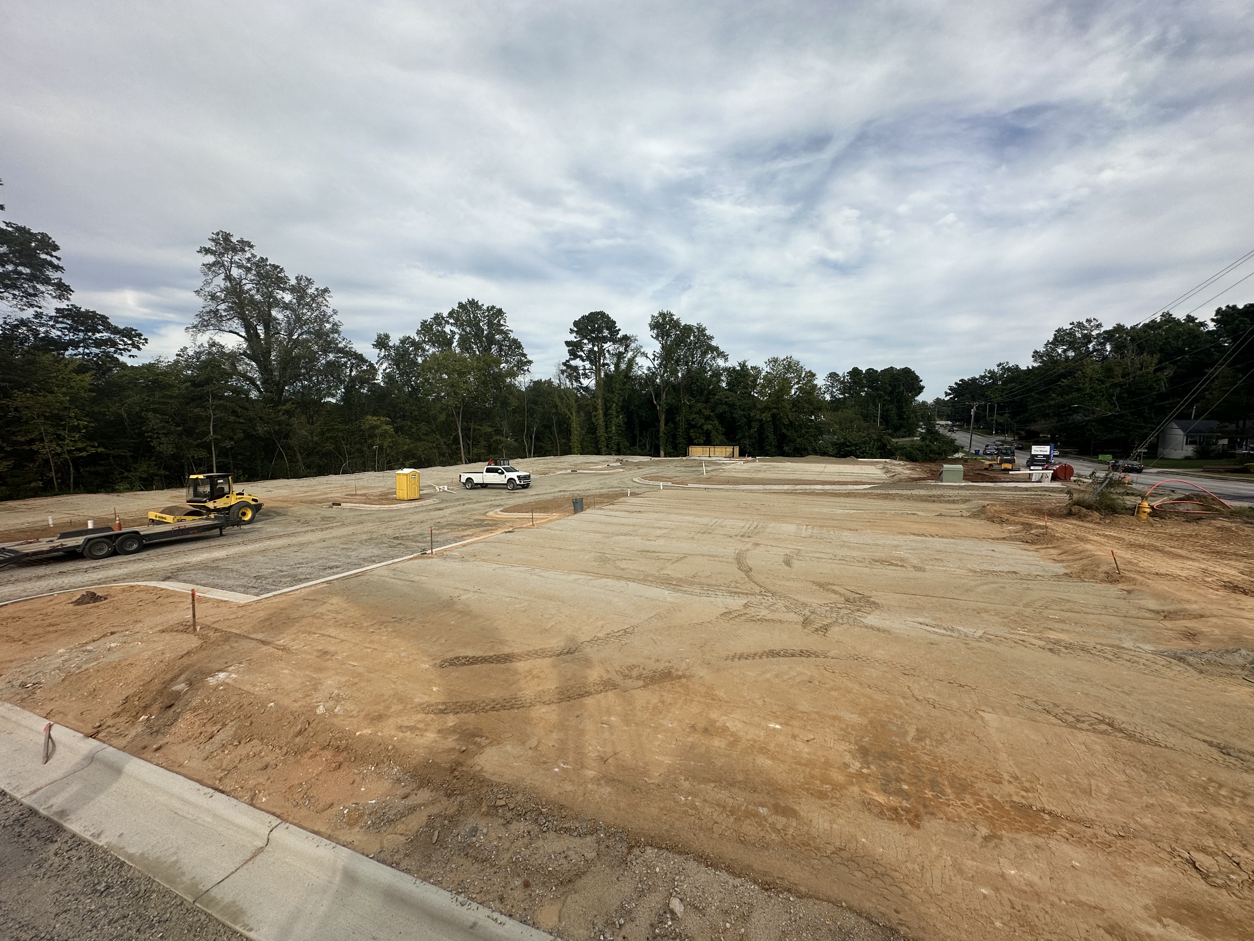 Construction site with cleared dirt areas, a white pickup truck, a yellow roller on a trailer, and trees in the background under a cloudy sky.
