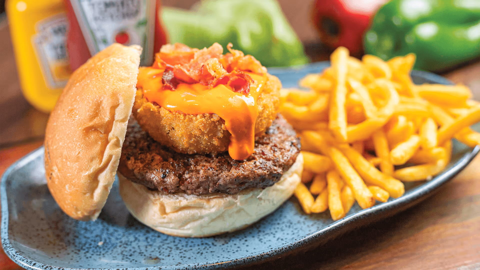 Hambúrguer com carne grelhada, queijo derretido, bacon crocante e anel de cebola empanado servido com batatas fritas em prato azul.