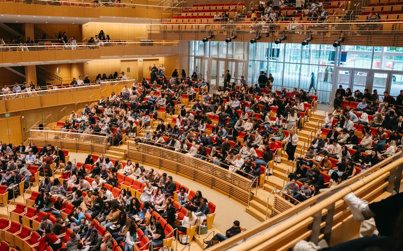 Large auditorium filled with people seated in tiered rows of orange chairs, with some standing and walking.