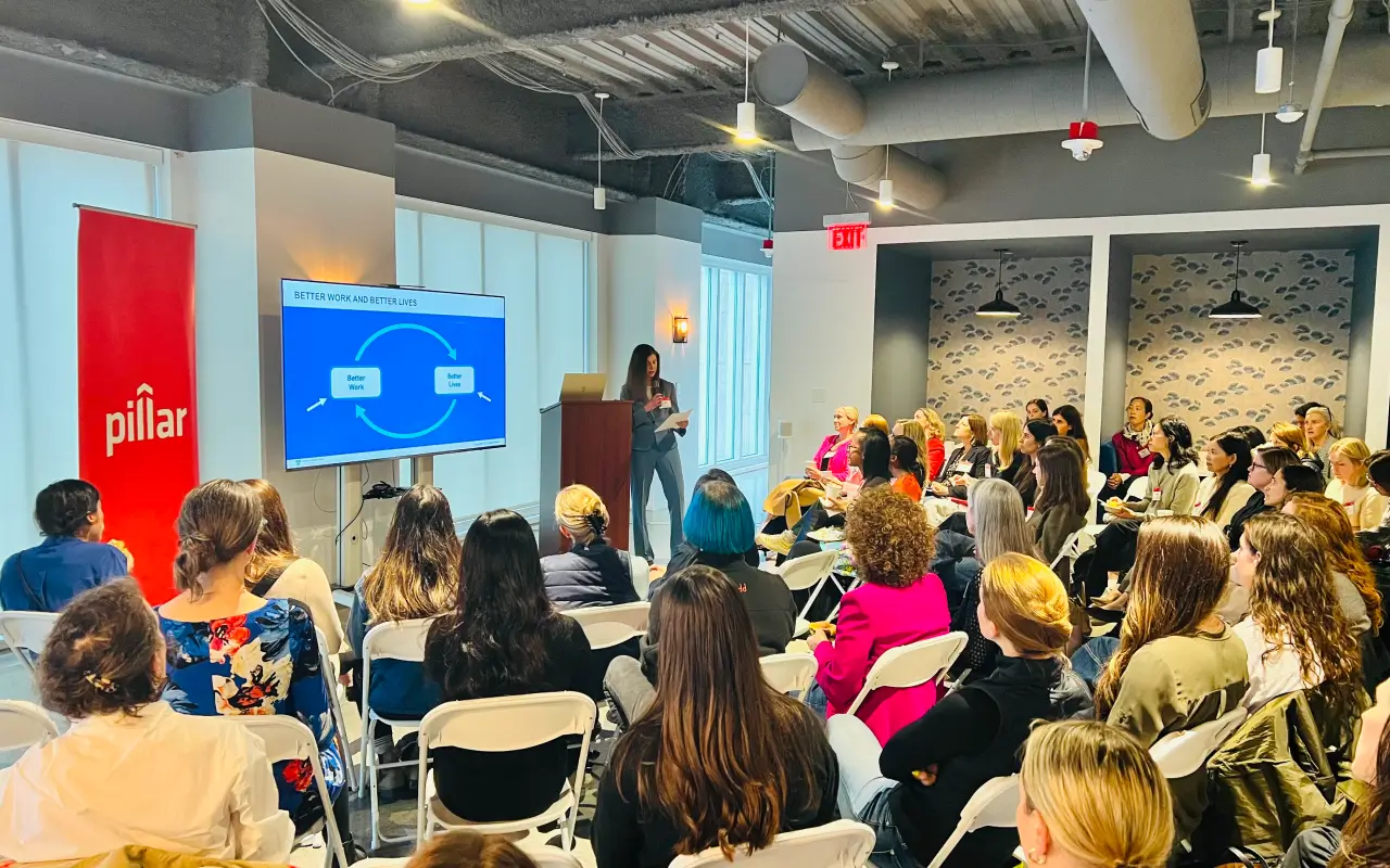Leslie giving a presentation to a seated audience in a modern conference room with a screen showing a cycle diagram labeled 'Better Work and Better Lives'.