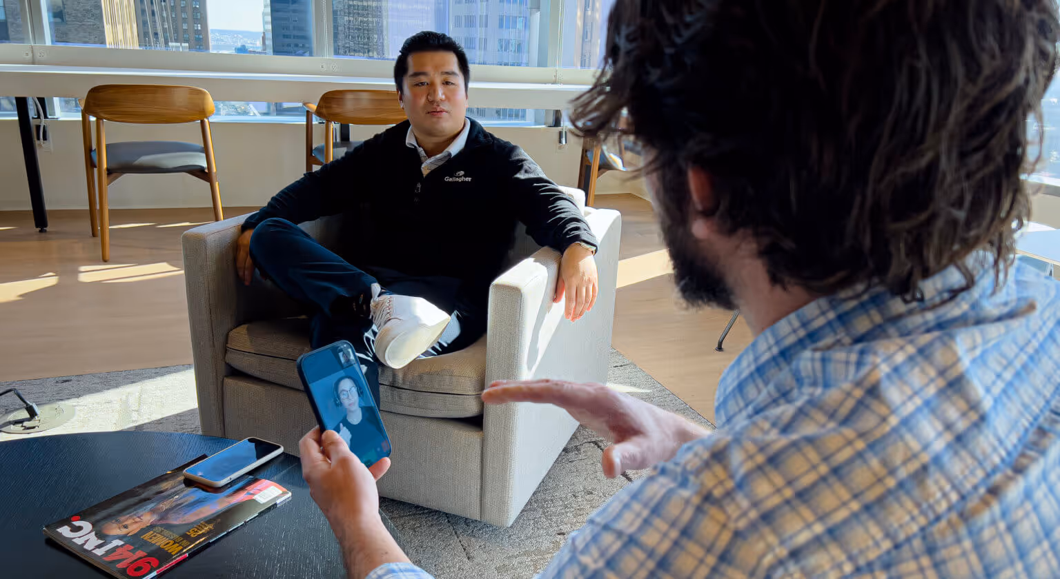 A modern office setting with large windows showcasing a cityscape. A man in a black sweater and white sneakers sits on a gray armchair, engaged in conversation with another person in a plaid shirt, who is holding a smartphone displaying a video call with a woman wearing glasses and a black top. A magazine titled 'Inc.' and another smartphone are placed on a black coffee table in the foreground.