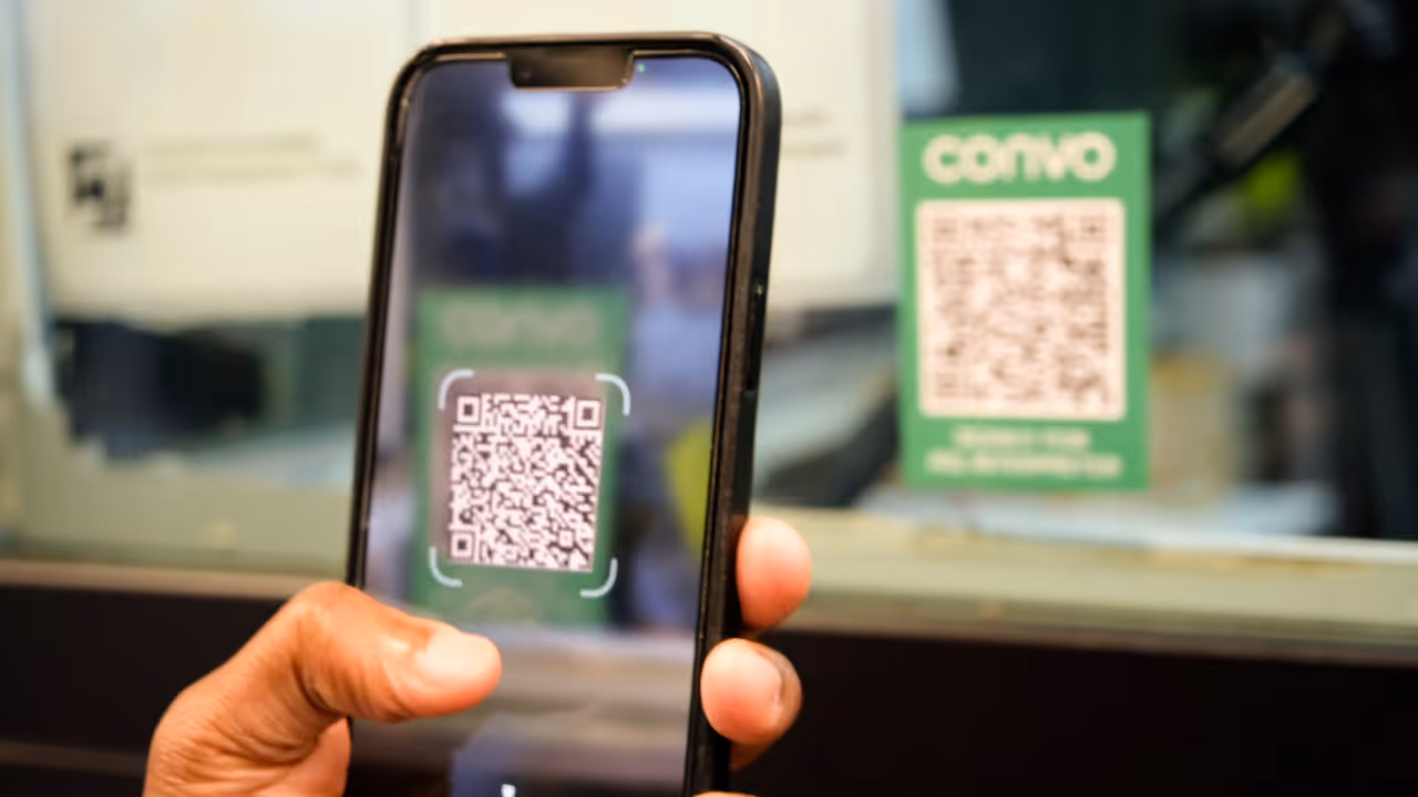 A close-up of a person holding a smartphone and scanning a QR code. The phone screen displays the QR code in focus, while the actual QR code is posted on a green sticker in the background with a logo 'Convo' in uppercase printed on it.