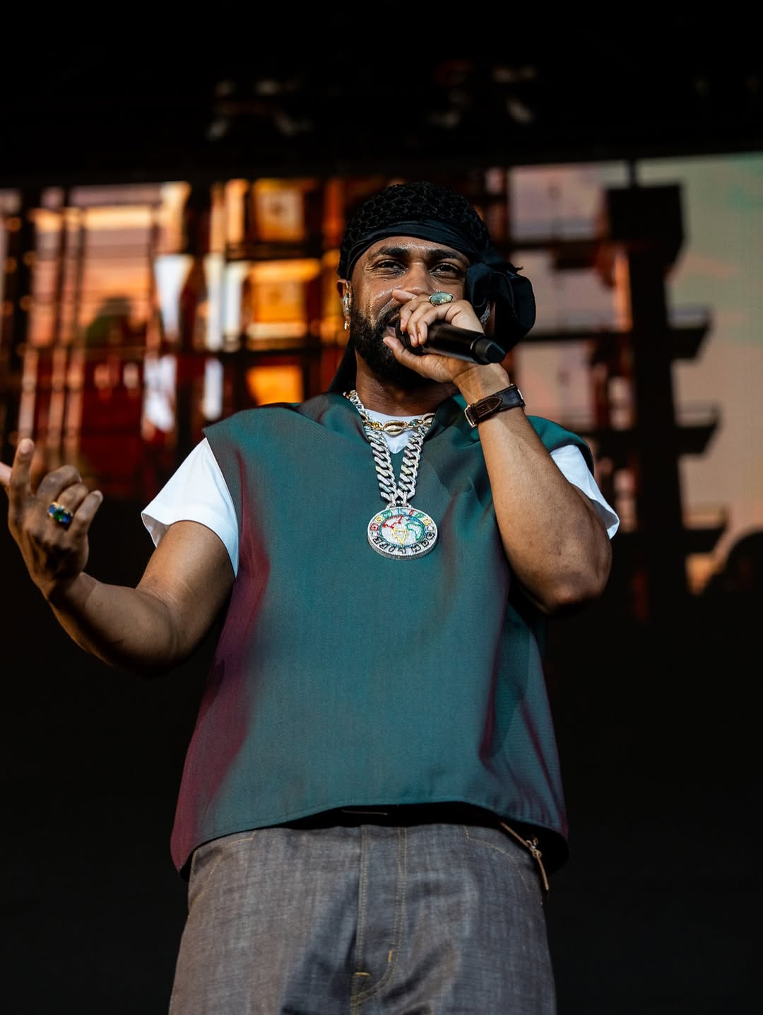 Rapper performing on stage with microphone, wearing teal vest and large necklace