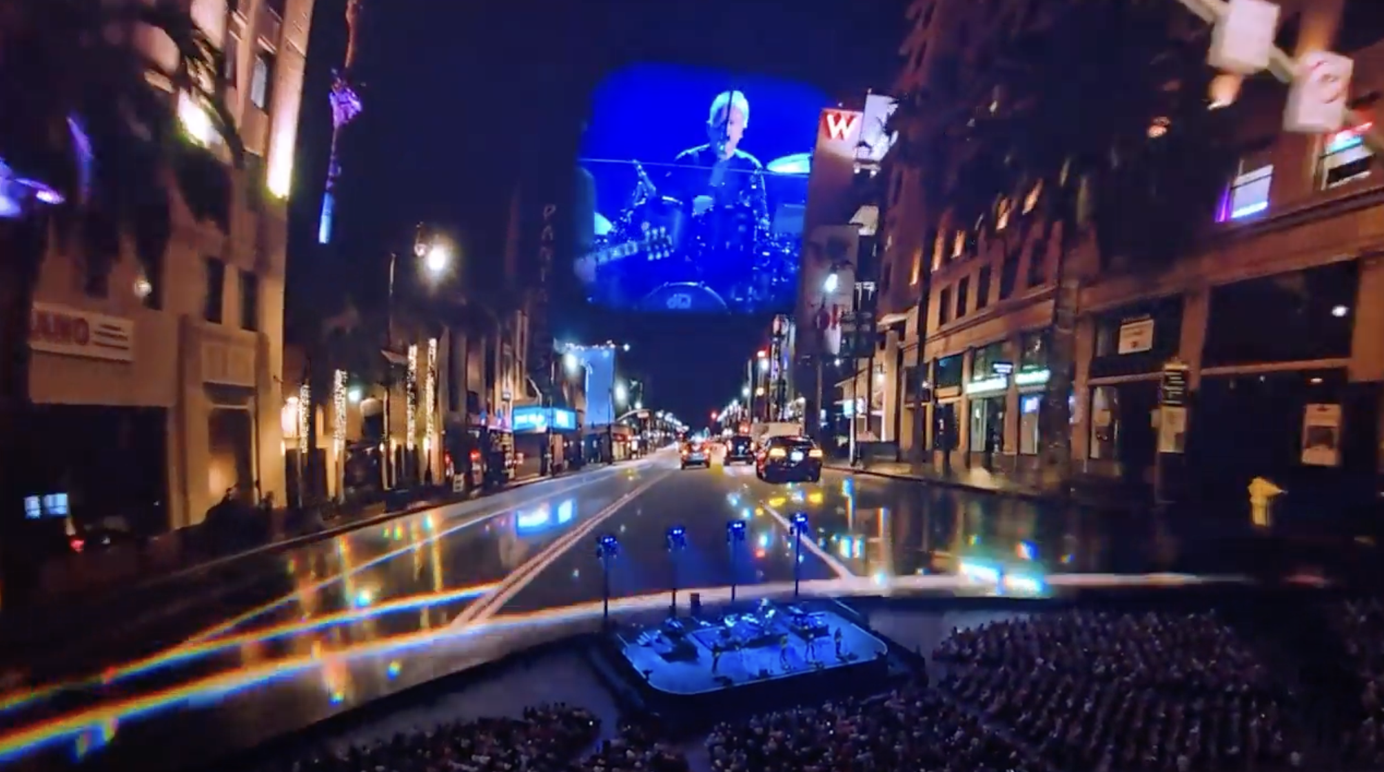 Large outdoor concert stage with drummer on screen in city street at night