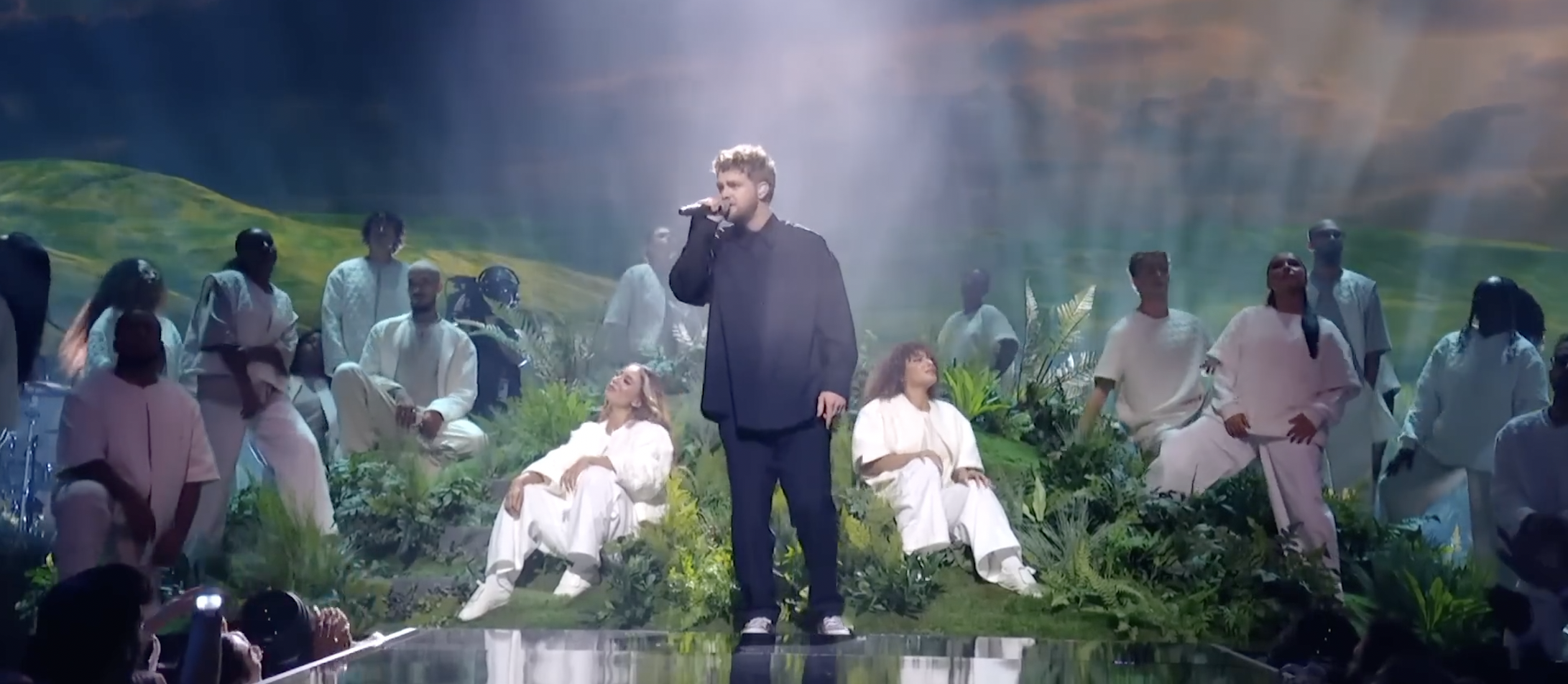 Singer performing on stage with dancers in white, surrounded by lush greenery